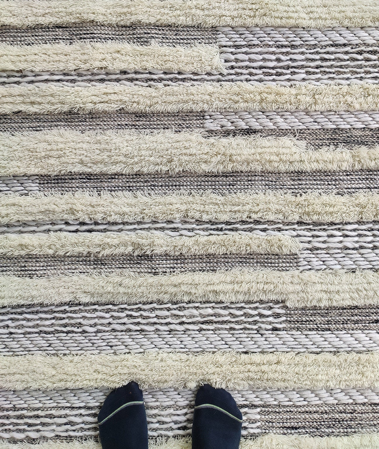 A pair of feet in black socks stands on the RAMSHA HOME Fringes Woven Rug SR-053, featuring beige, cream, and brown stripes with various woven patterns and shaggy textured sections—perfect for living rooms or bedrooms.