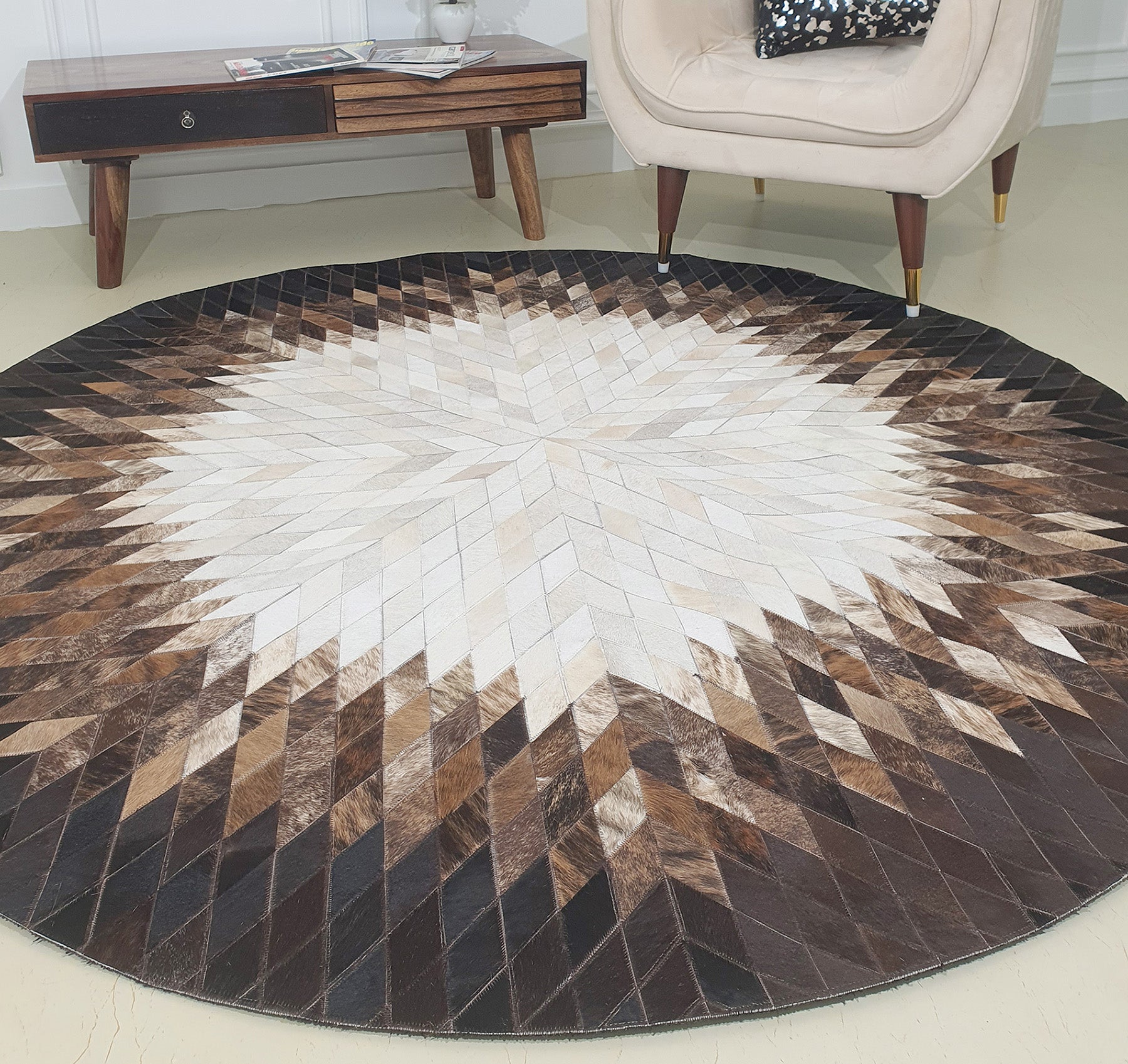 The RAMSHA HOME Multi Color Home Decor Round Leather Rug - LR 017 features a geometric starburst pattern in brown, beige, and white. It's shown on a light floor near a cream armchair and wooden side table.