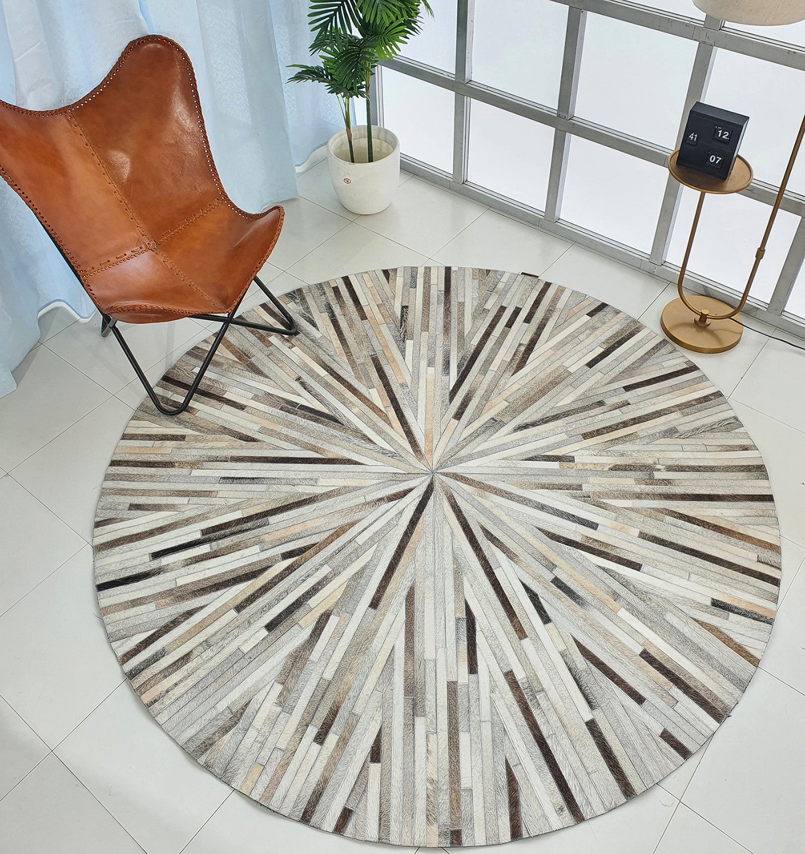 A modern living space featuring the RAMSHA HOME Home Decorative Leather Rug - LR 018 with a sunburst pattern, paired with a brown butterfly chair, potted plant, side table, and digital clock near large windows.