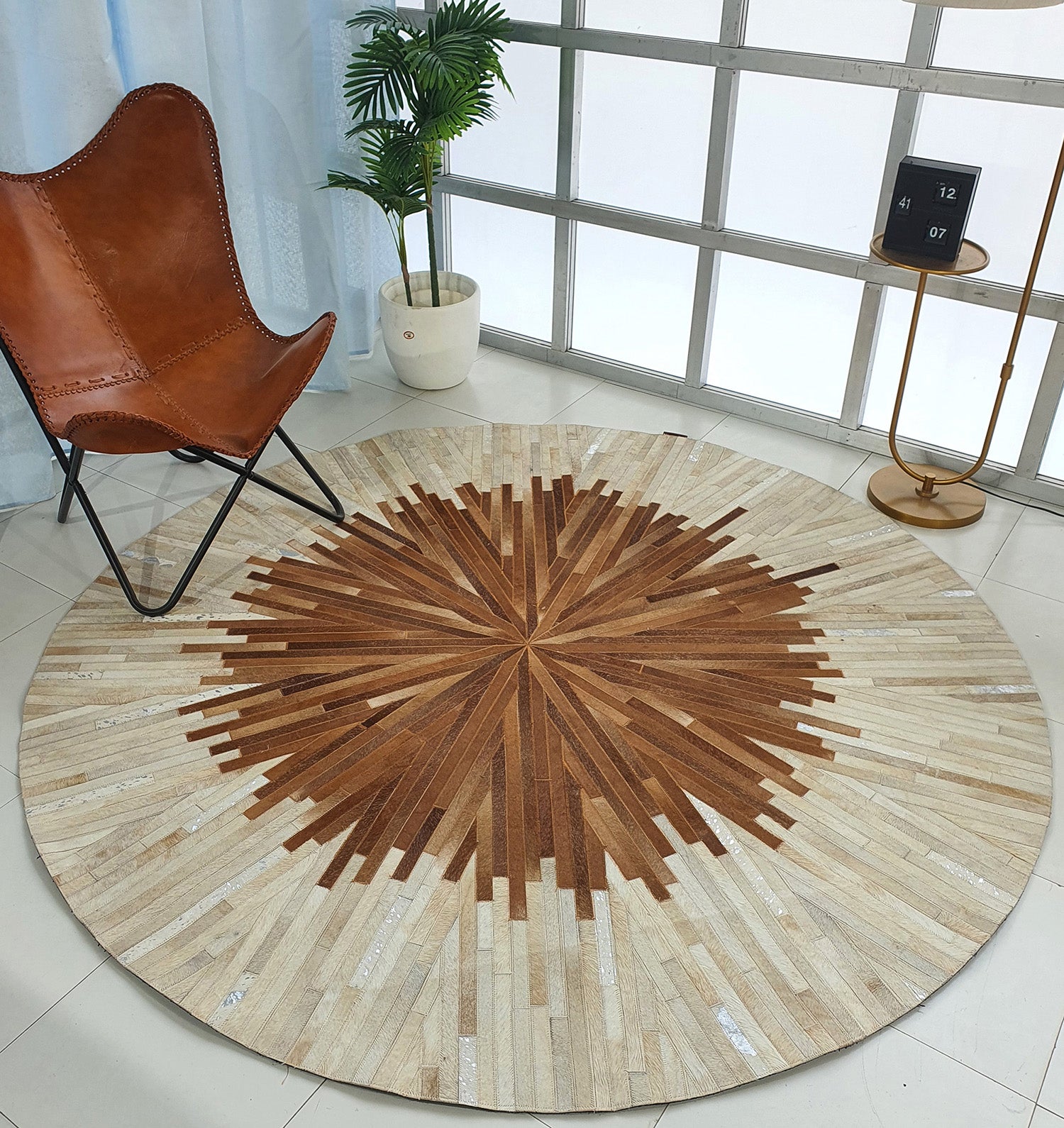 The Multi Color Round Leather Rug - LR 016 features a geometric sunburst design in brown and beige tones, displayed on a tiled floor beside a brown leather chair, tall plant in a white pot, and gold floor lamp with a black calendar.