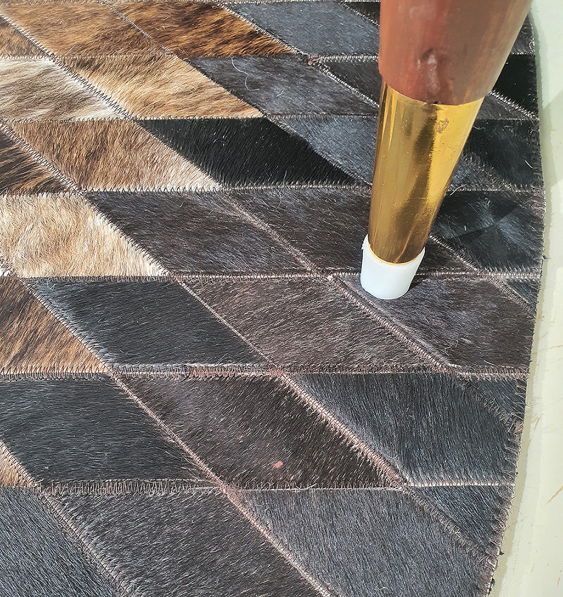A close-up of RAMSHA HOME’s Multi Color Home Decor Round Leather Rug - LR 017 displays its geometric leather patchwork with tan fur accents, while a gold and brown furniture leg with a white tip sits atop the detailed stitching.
