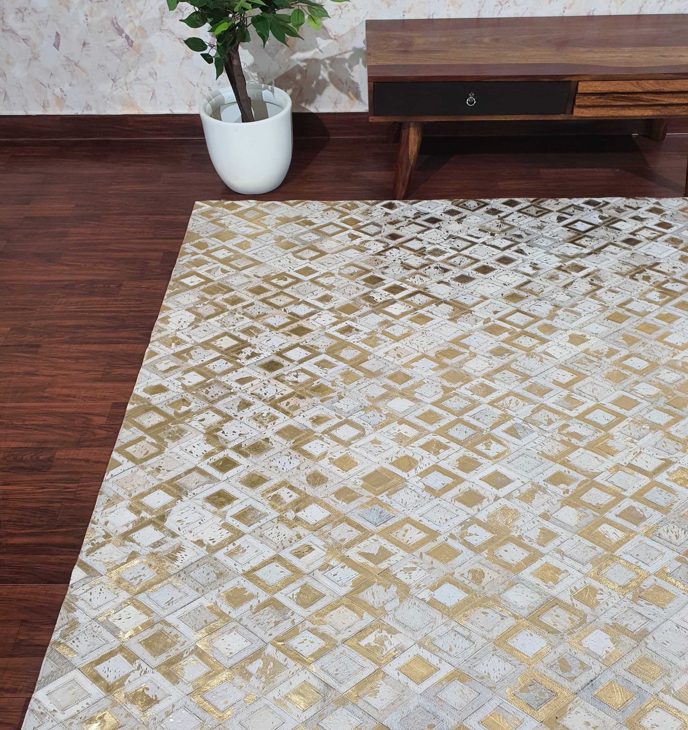 A modern living room corner with a wooden floor, a RAMSHA HOME Handmade Diamond Patterned Leather Rug LR-010 in white and gold, a white potted plant, and a low wooden table with a drawer against a light-colored wall.