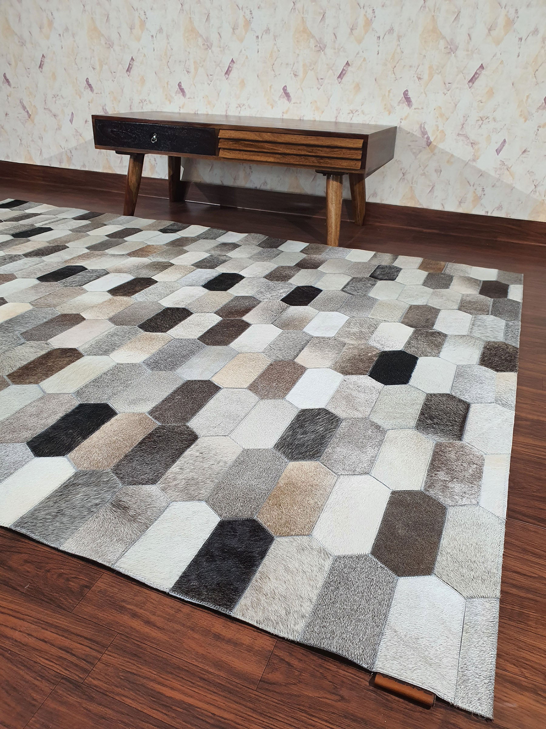 The RAMSHA HOME Multi Color Home Decor Leather Rug - LR 011, featuring hexagonal and rectangular shapes in neutral tones, rests on a dark wood floor beside a mid-century modern table and a light textured wall.