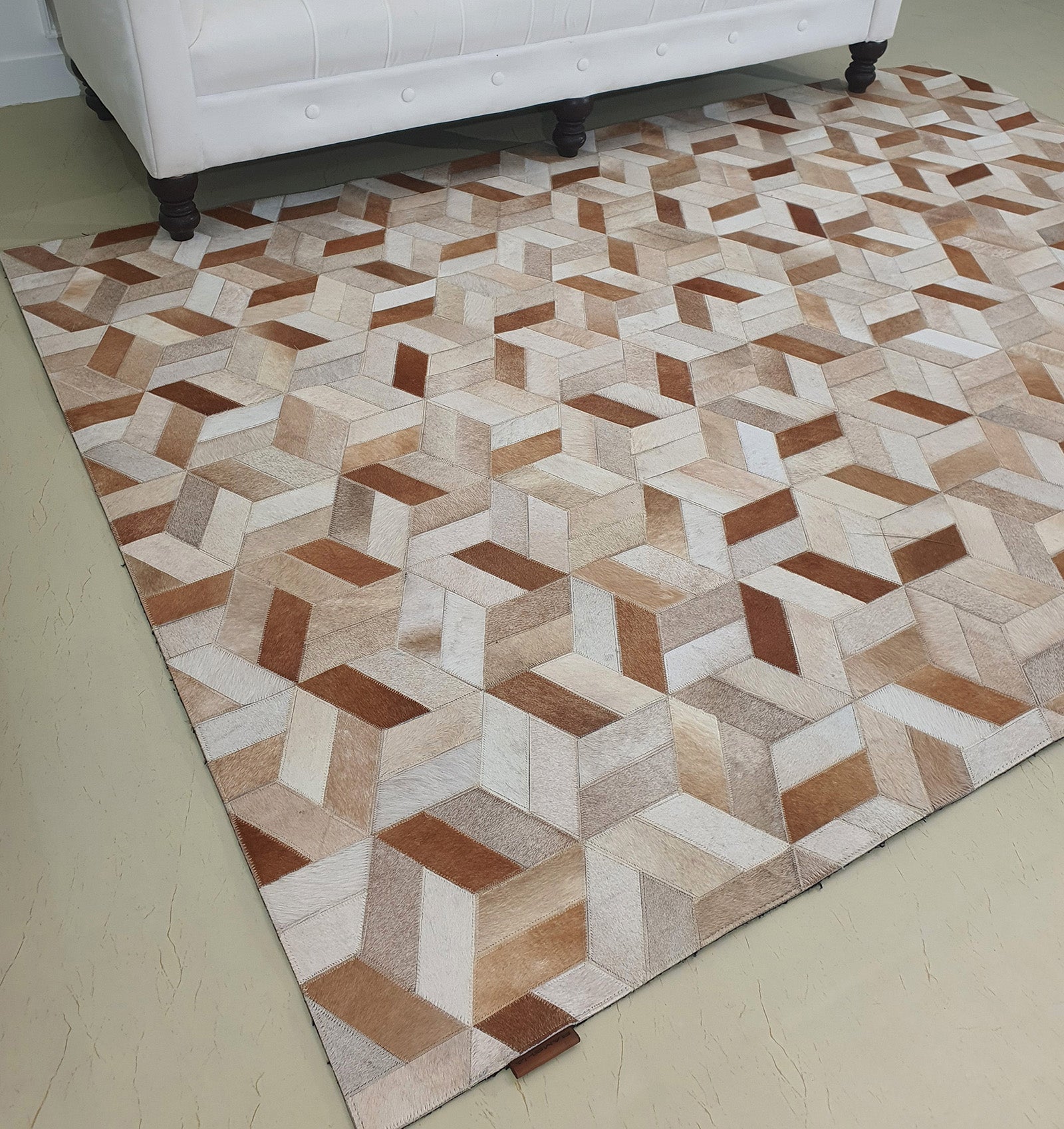 The RAMSHA HOME Geometric Cube Pattern Handmade Leather Area Rug LR-012, featuring tan, beige, brown, and cream cubes for a 3D effect, is displayed on a light floor before a white upholstered sofa with dark wooden legs.