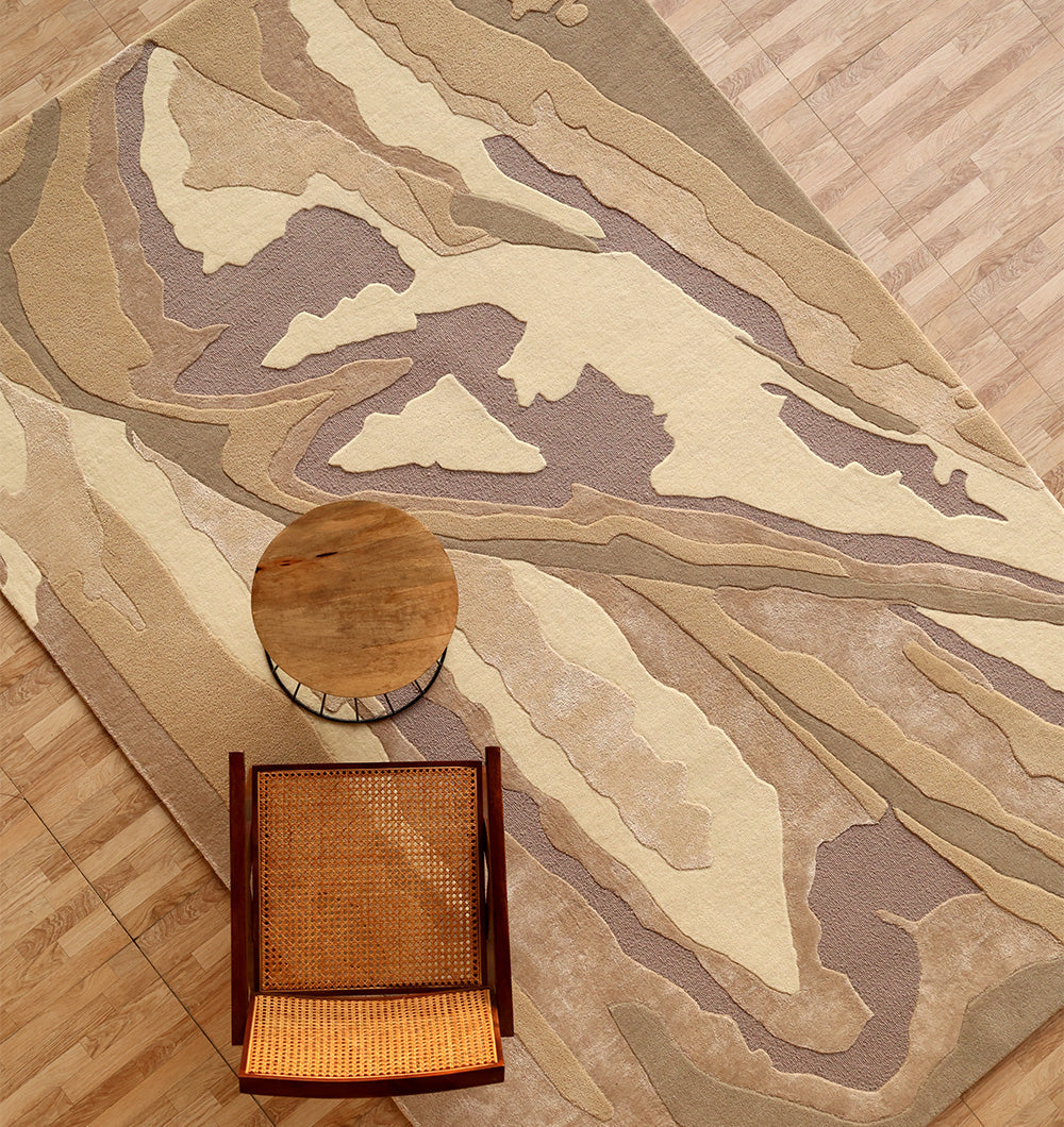 A top-down view of the RAMSHA HOME Abstract Wool & Viscose Handmade Carpet 200x300 (HTWV-179), featuring earth-toned geometric patterns, with a round wooden side table and woven cane chair on a light wood floor.