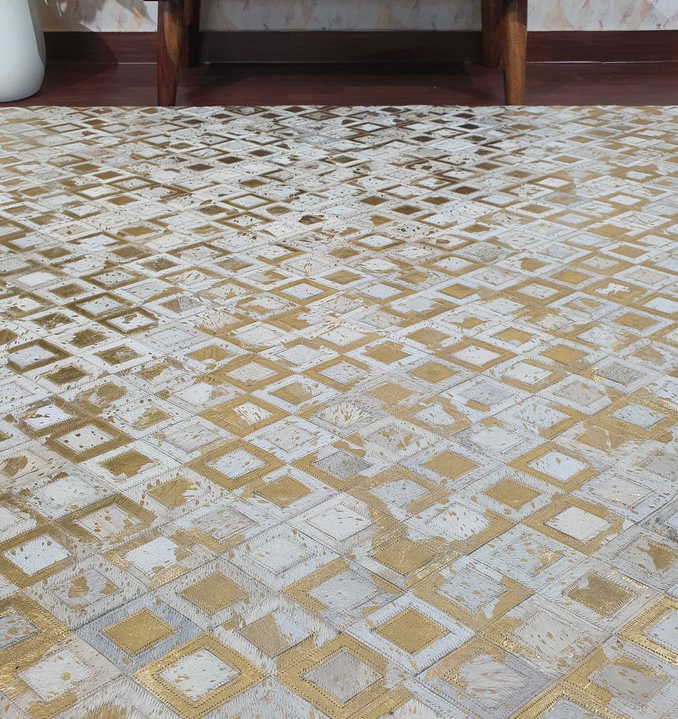 A close-up of the RAMSHA HOME Handmade Diamond Patterned Leather Rug LR-010, featuring beige, white, and gold squares and diamonds, displayed on a wooden floor by a wall.