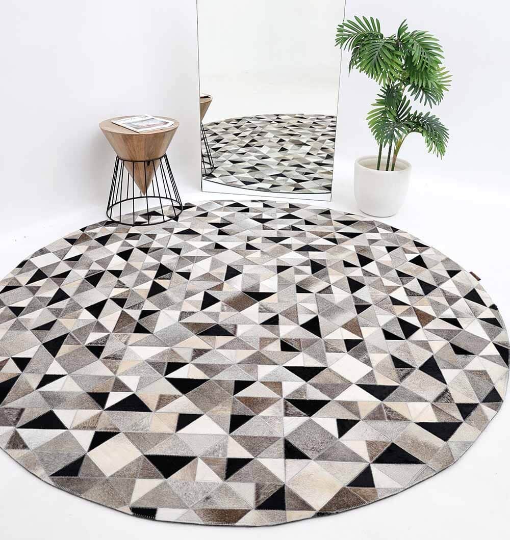 The RAMSHA HOME Multi Color Home Decor Leather Rug - LR 020 featuring black, white, and gray triangles lies on the floor beside a small wooden side table with a magazine, a potted plant, and a tall mirror reflecting the decor.