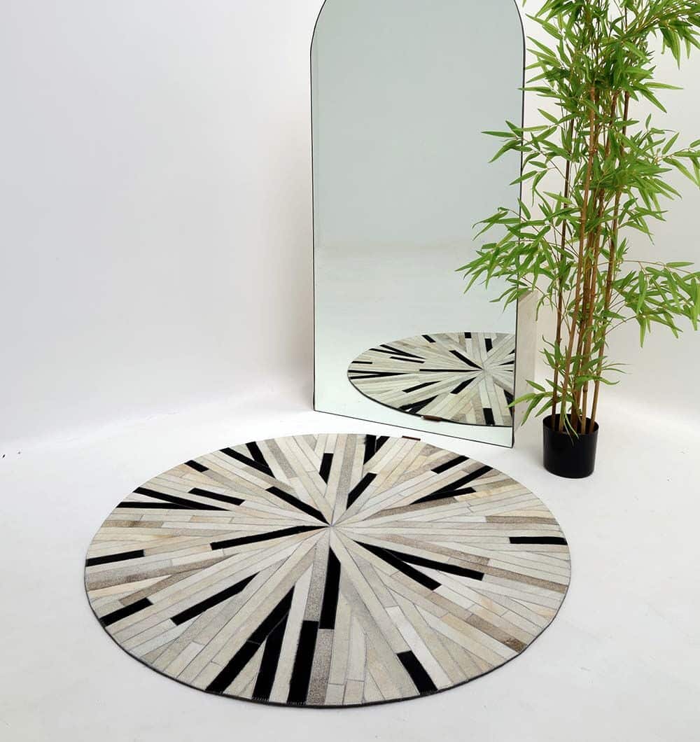 The RAMSHA HOME Stunning Multi Color Dining Room Decor Leather Rug - LR 021, featuring a round design with geometric sunburst patterns in beige, white, and black, is placed on the floor next to a tall arched mirror and a potted plant with slender green leaves.