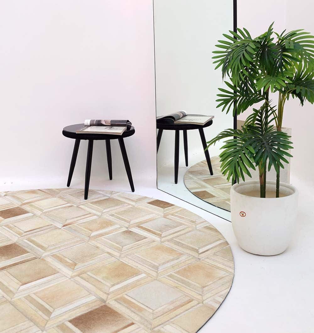 The Multi Color Round Leather Rug - LR 023 by RAMSHA HOME, a black round side table with a book, and a potted plant are placed in front of a large mirror in a minimalist white-walled room.