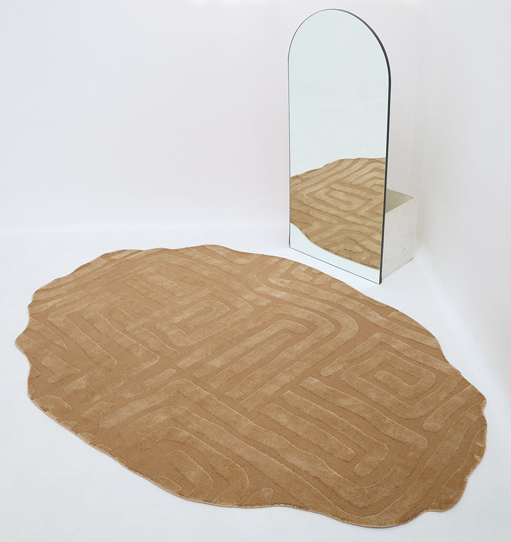 The Amazing Brown Color Unshape Decor Rug (200x300cm) by RAMSHA HOME, featuring a geometric design, lies on a white floor beside an arched mirror with a white base that reflects the rug’s modern style.