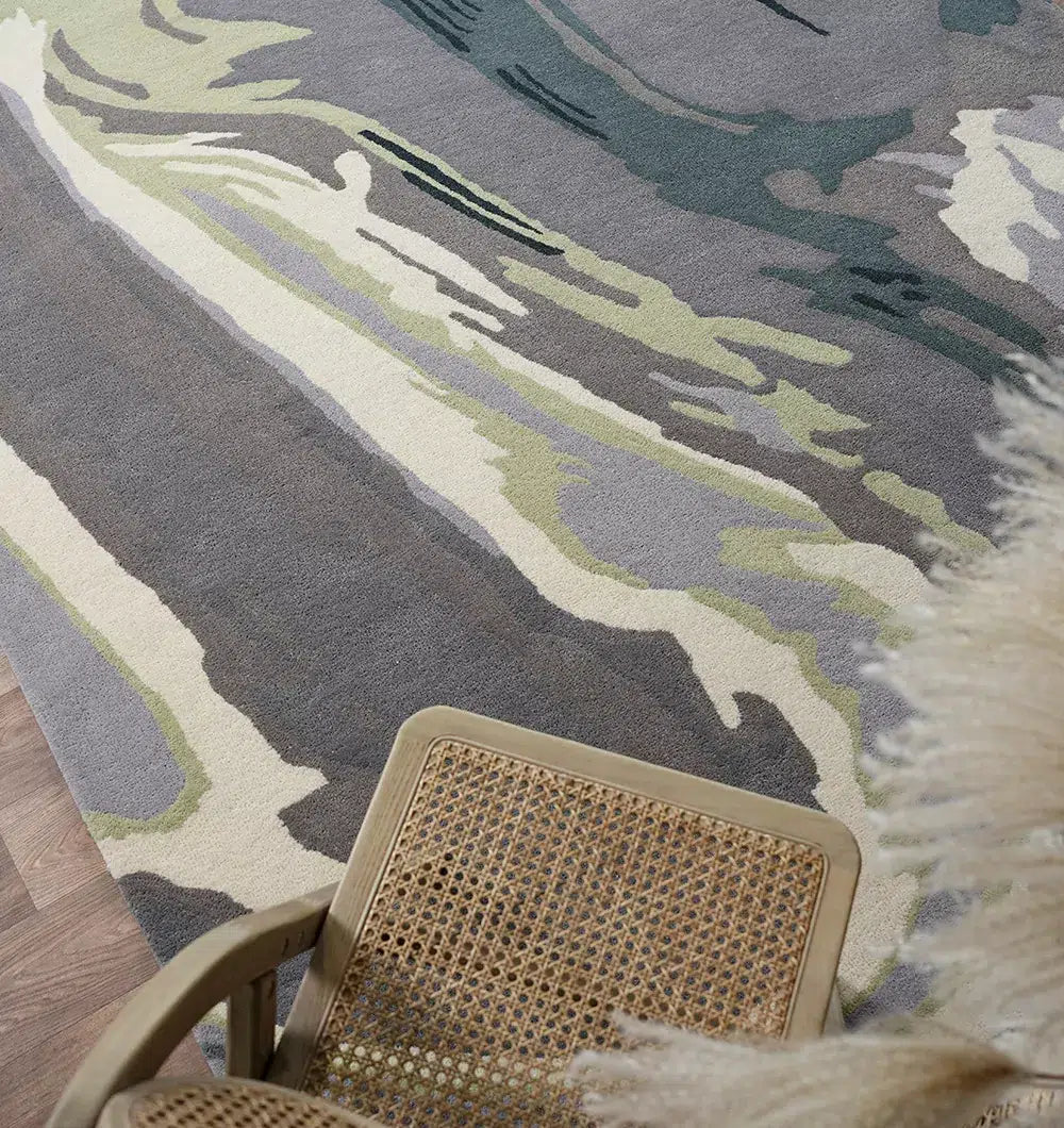 A woven chair with a cane seat and a cream fur throw sits beside the RAMSHA HOME Beautiful Hand-Tufted Rug - CWL 11 (200x300cm), featuring grey, beige, cream, and green tones, on a wooden floor.