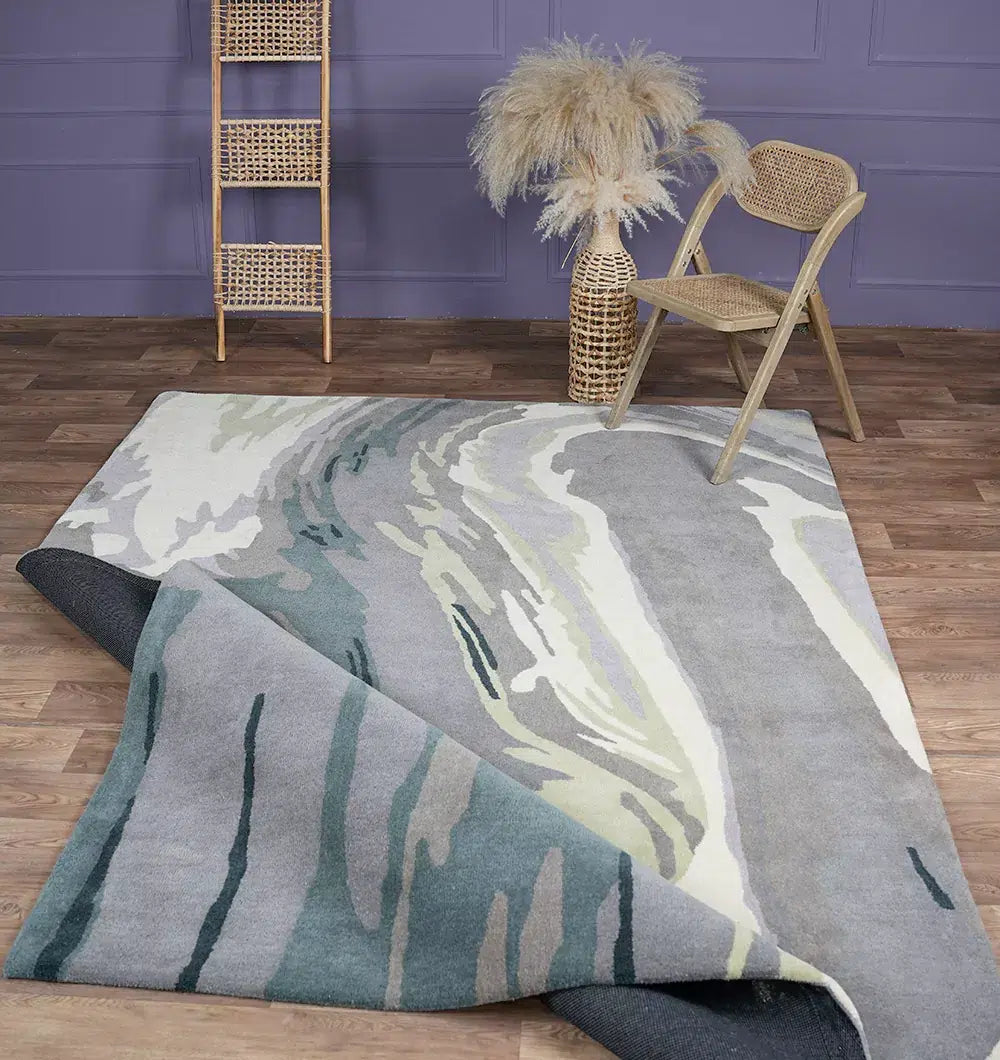 A modern room featuring the RAMSHA HOME Beautiful Hand-Tufted Rug - CWL 11 (200x300cm) in blue-grey wool, partially folded, with a rattan chair, ladder shelf, and tall vase with pampas grass against a purple wall and wood floor.