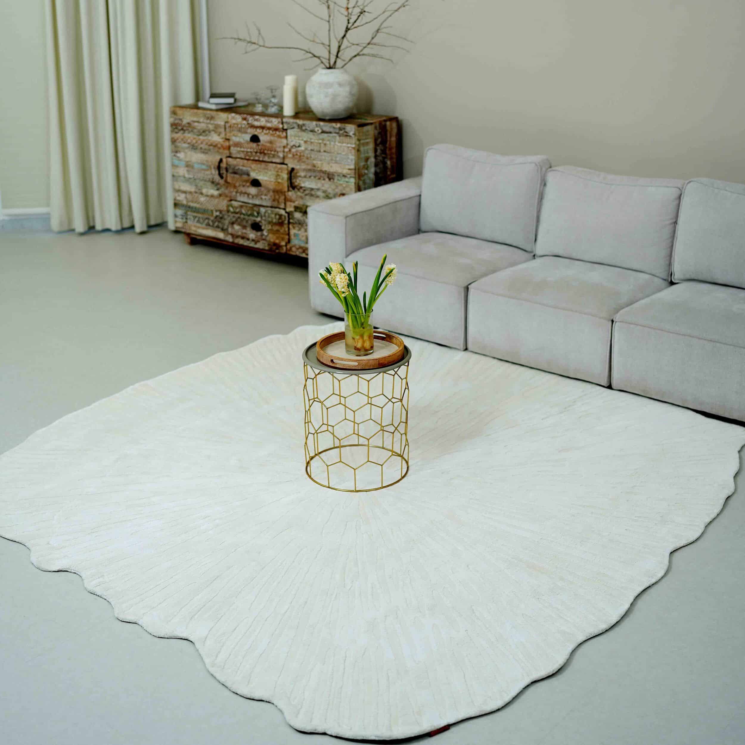 A minimalist living room with a light gray couch, rustic wooden cabinet, round coffee table with a gold geometric base, and the RAMSHA HOME Home Decor Shape Handmade Area Rug - HTV 21. The table holds a vase of white flowers.
