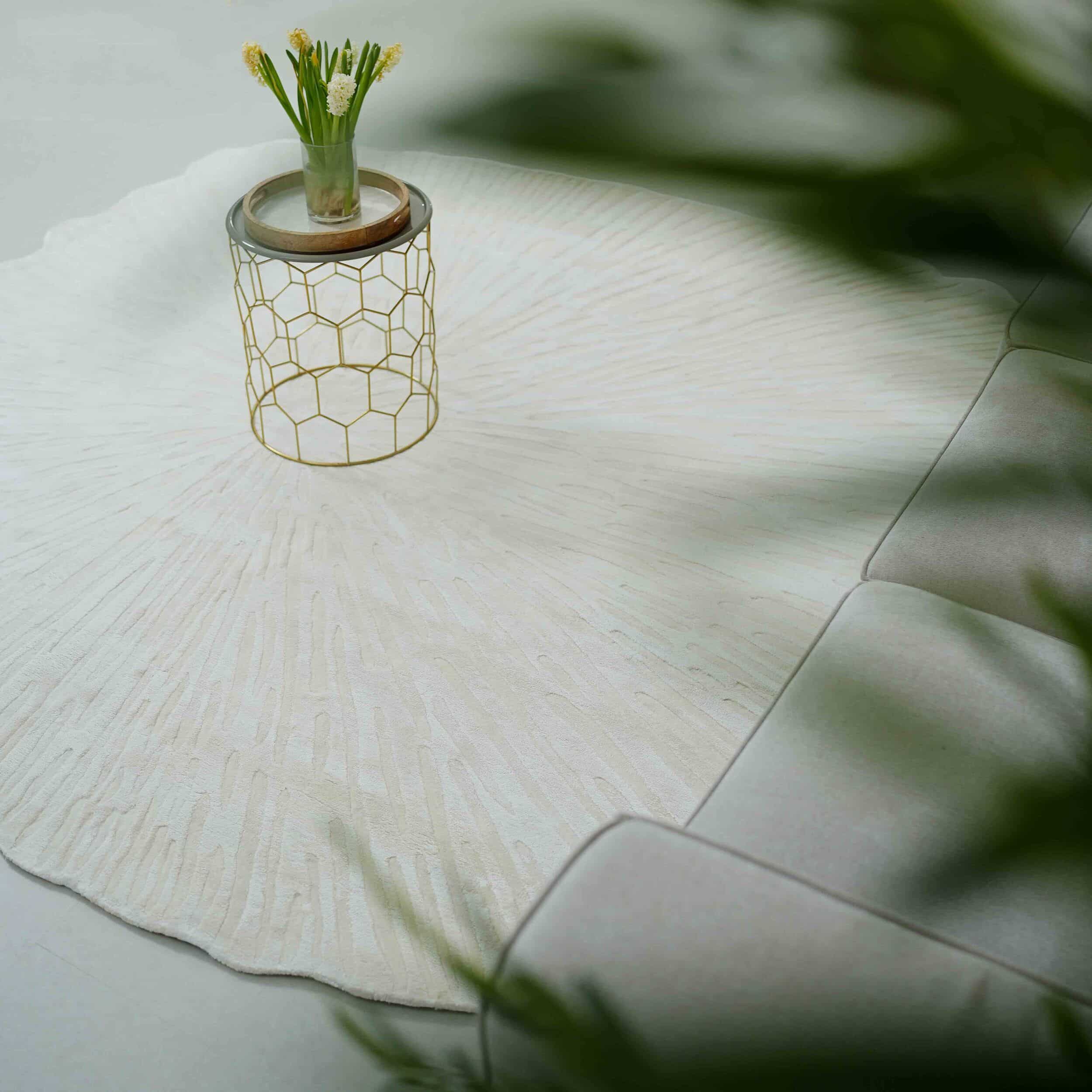 A modern living space featuring the RAMSHA HOME Home Decor Shape Handmade Area Rug - HTV 21 in ivory, a metallic honeycomb side table with yellow flowers, and part of a light gray sofa. Blurred green leaves appear in the foreground.