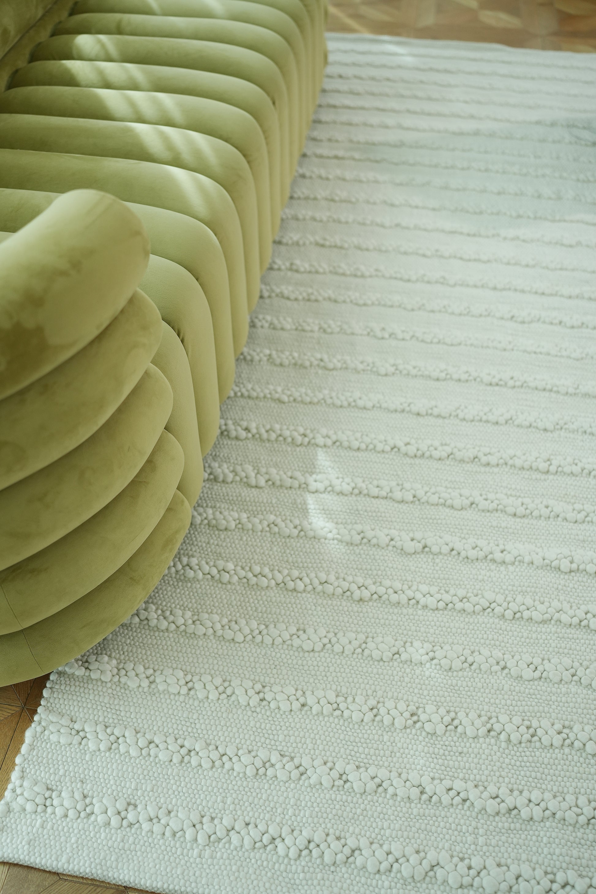 A close-up of a light green curved sofa beside the RAMSHA HOME Home Decor Woven Rug Natural Color Bedroom Rug SRN-037 on a wooden floor, with sunlight casting soft shadows across the scene.