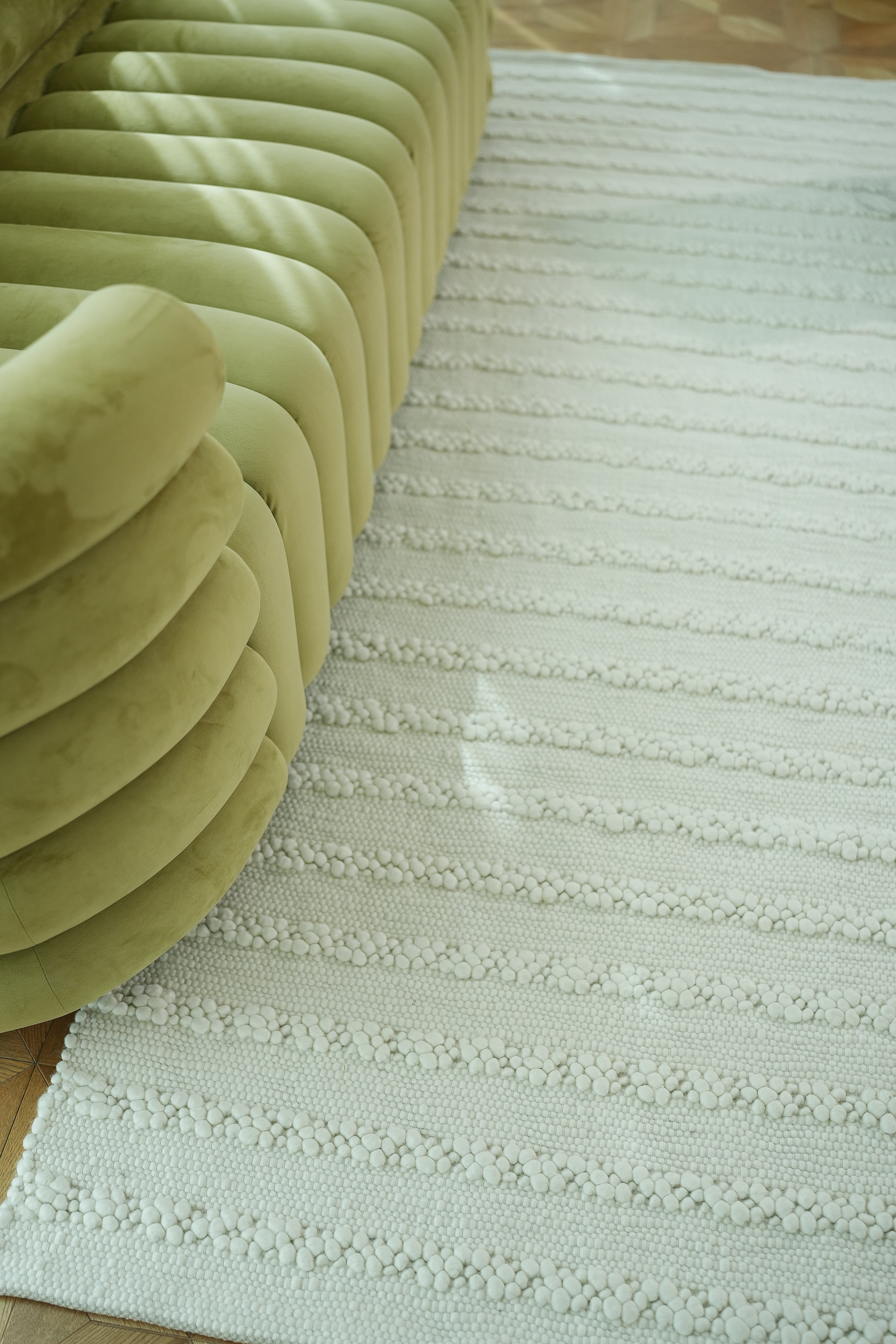 A close-up of a light green curved sofa beside the RAMSHA HOME Home Decor Woven Rug Natural Color Bedroom Rug SRN-037 on a wooden floor, with sunlight casting soft shadows across the scene.