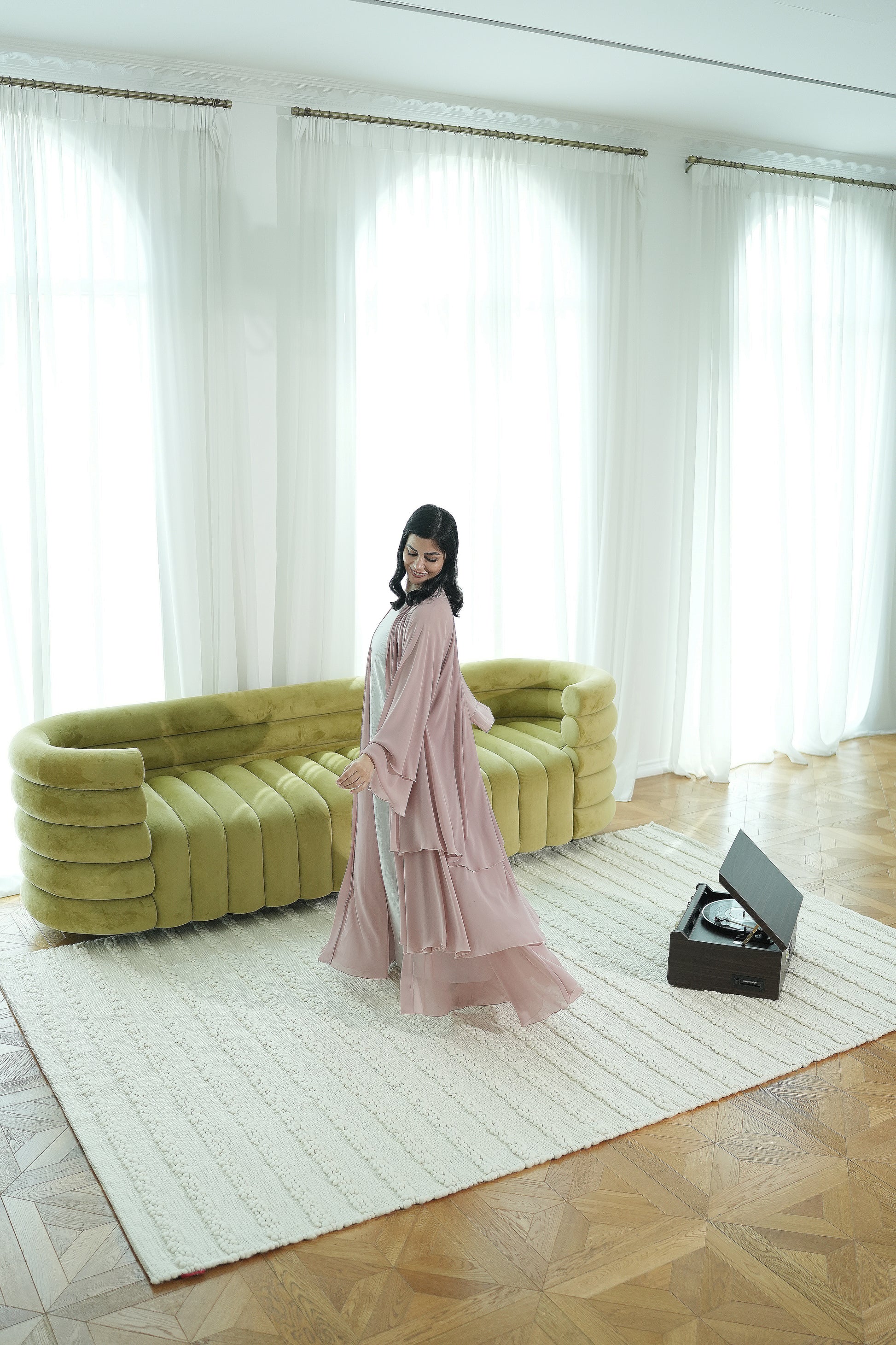 A woman in a flowing pink dress stands on the RAMSHA HOME Home Decor Woven Rug Natural Color Bedroom Rug SRN-037 in front of a green sofa, with sheer white curtains behind her and an open record player to her right.