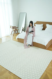 A woman in a light pink robe reads on a neat bed in a bright, modern bedroom with a large mirror and wooden desk. The RAMSHA HOME Home Decor Woven Rug Natural Color Area Rug SRN-038 adds warmth to the inviting space.