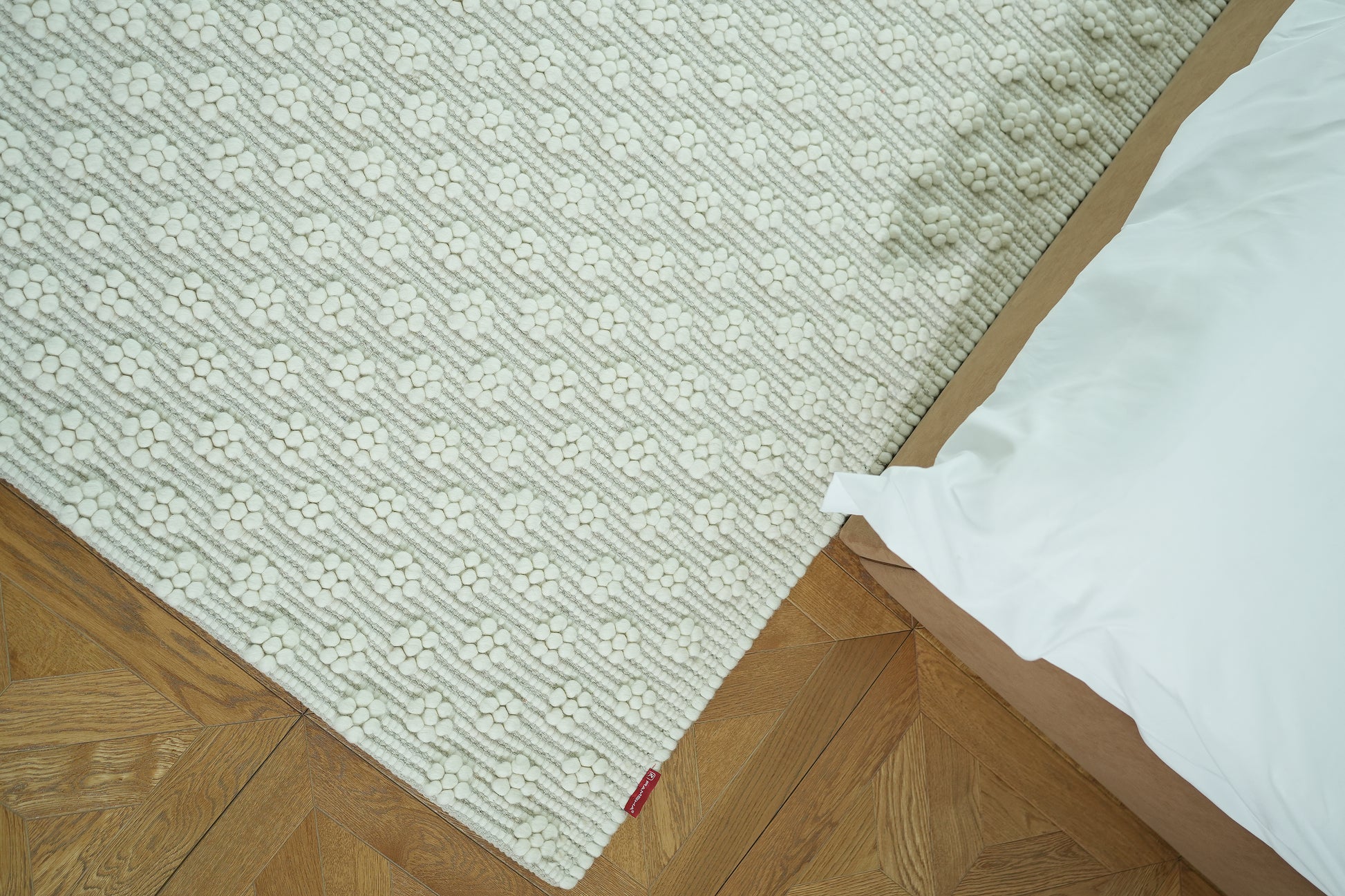 The RAMSHA HOME Home Decor Woven Rug Natural Color Area Rug SRN-038, featuring a geometric pattern, is placed on a wooden parquet floor by a bed with white bedding. A stained spot is visible near the edge of this neutral rug.