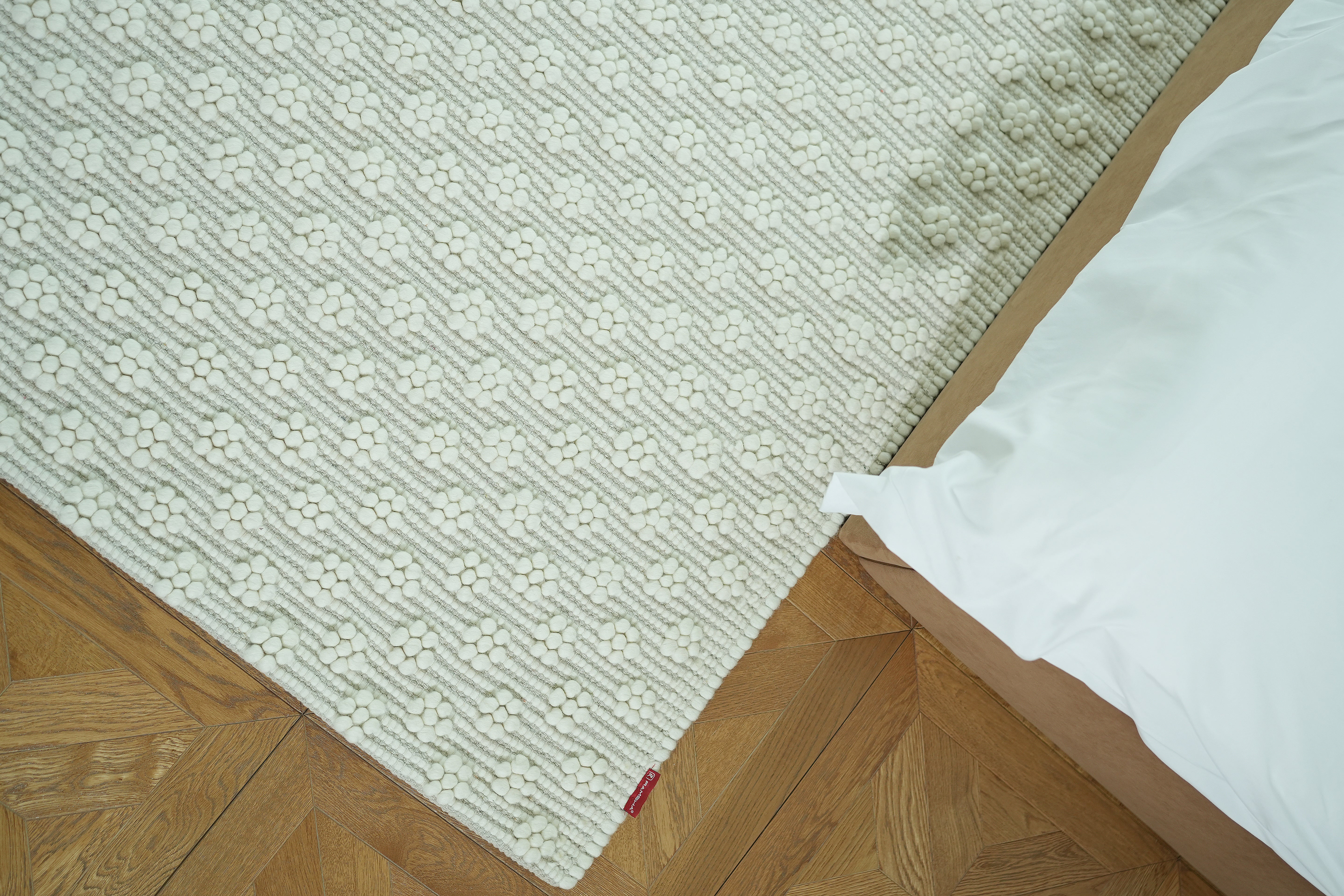 The RAMSHA HOME Home Decor Woven Rug Natural Color Area Rug SRN-038, featuring a geometric pattern, is placed on a wooden parquet floor by a bed with white bedding. A stained spot is visible near the edge of this neutral rug.