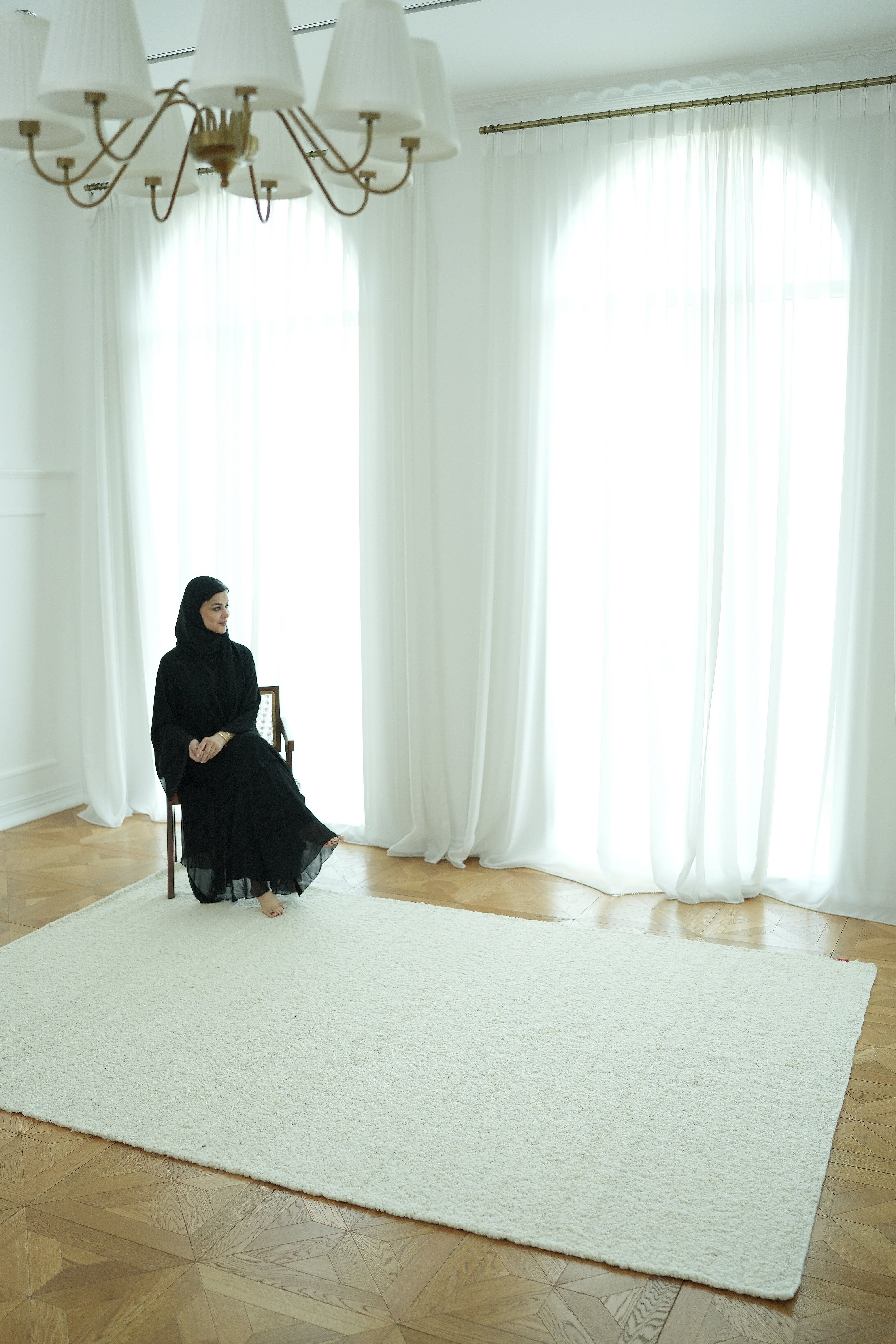 A woman in a black dress and hijab sits on a chair in a bright, minimalist room featuring sheer white curtains, a chandelier, wooden floor, and the RAMSHA HOME Handmade Natural Off White Woven Rug For Home Decor SR-059.