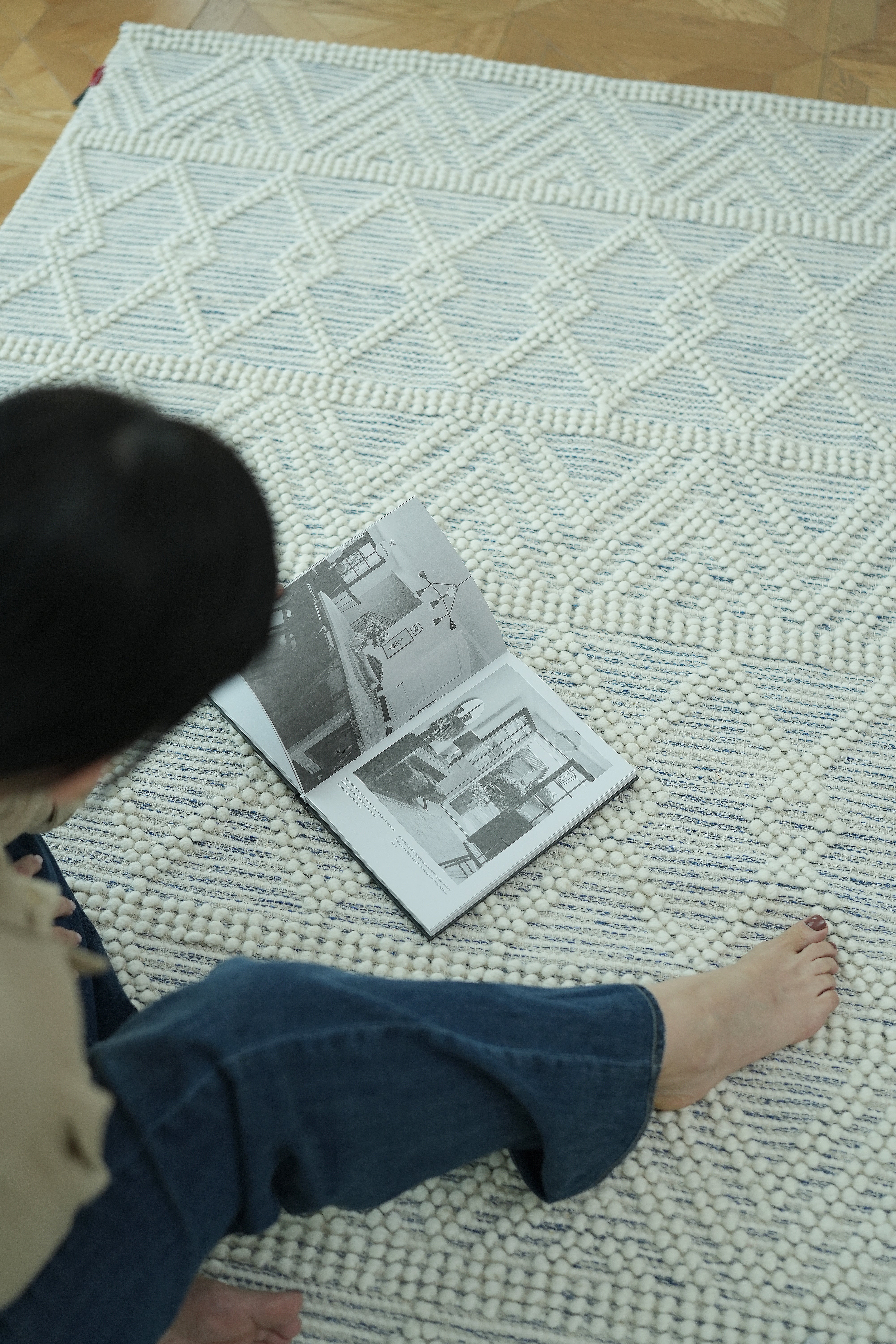 A person sits barefoot on the RAMSHA HOME Handmade Woven Rug Natural & White Color Floor Rug SRN-021, looking at an open book with black-and-white interior photos. The person's face is not visible.