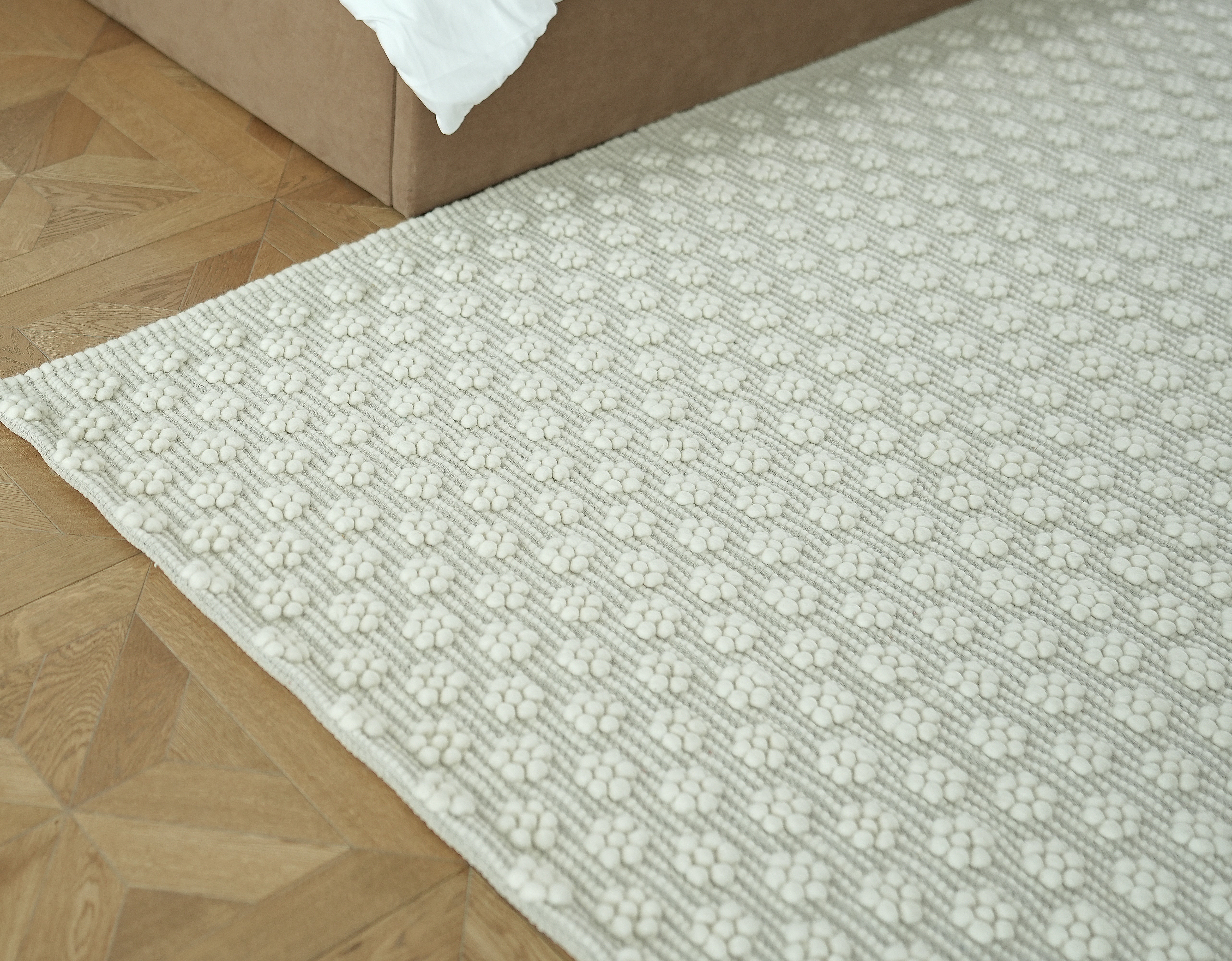 A close-up of RAMSHA HOME's Home Decor Woven Rug Natural Color Area Rug SRN-038, featuring a geometric texture, lies on a light wooden floor beside a beige bed frame with white bedding, highlighting the rug’s neutral charm.