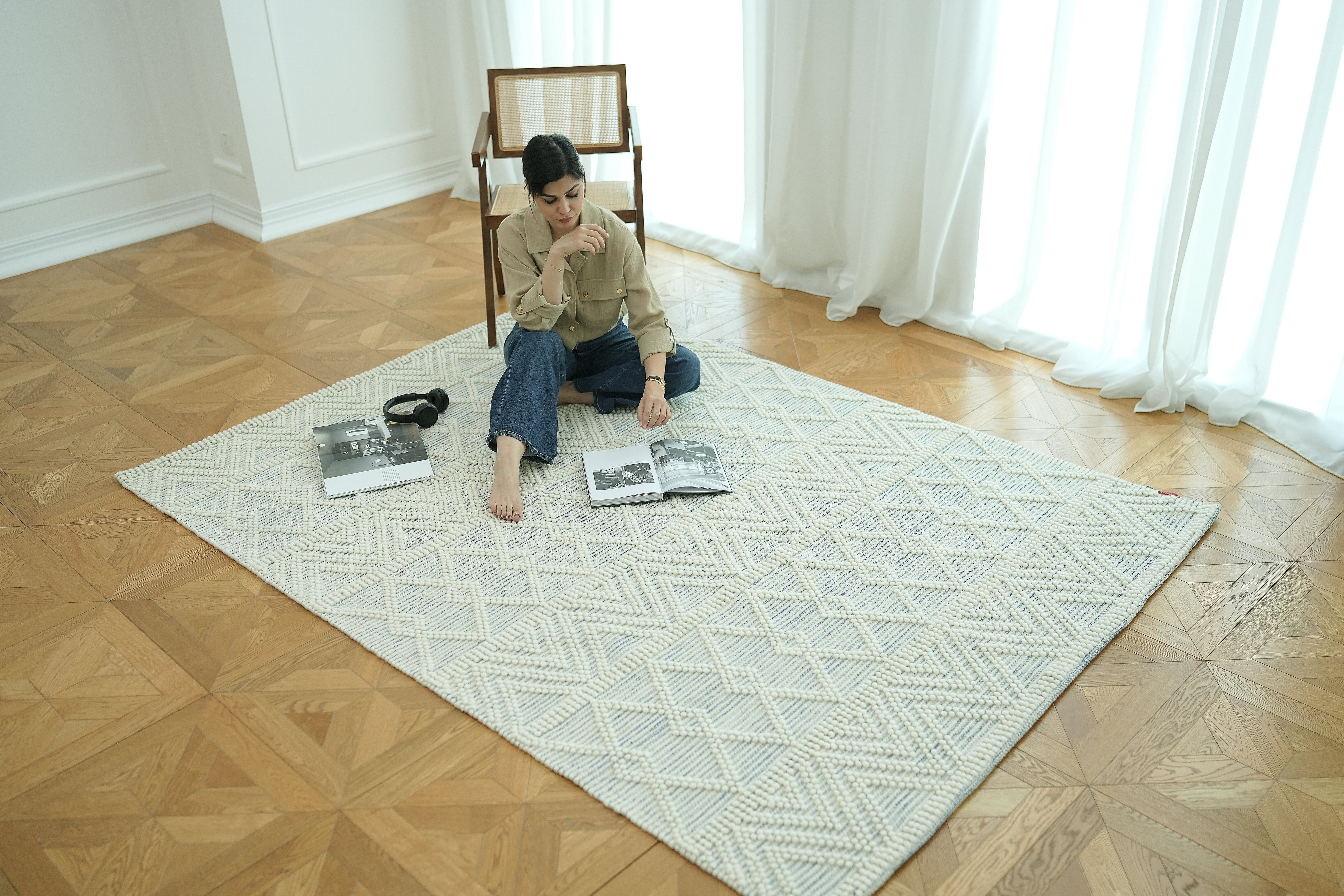 A person reads a magazine while sitting cross-legged on the RAMSHA HOME Handmade Woven Rug SRN-021 in natural and white, in a bright room with wooden floors. Nearby are a chair, headphones, another magazine, and large windows with white curtains.