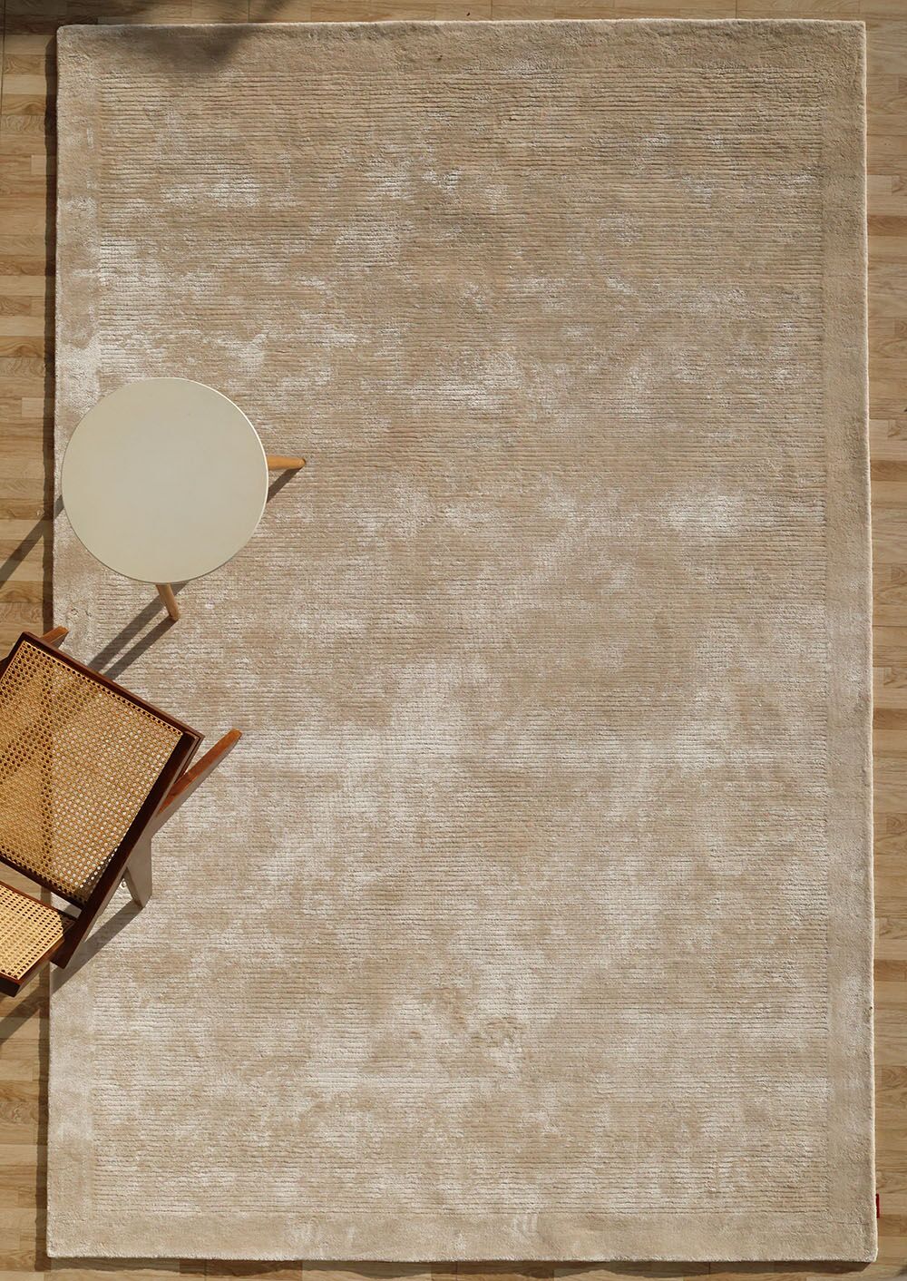 A RAMSHA HOME Beige Color Handmade Rug - HTV 18 (200x300 cm) is styled on a light wood floor, paired with a light round side table and a woven chair in the bottom left corner, captured from an overhead view.
