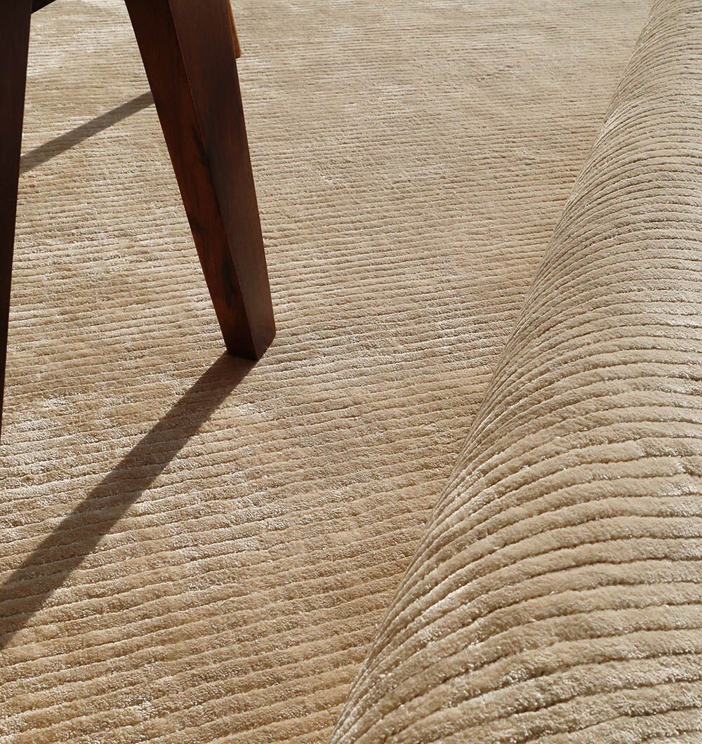 A close-up of the RAMSHA HOME Beige Color Handmade Rug - HTV 18 (200x300 cm) being unrolled beside a wooden furniture leg, highlighting its textured, ribbed surface.