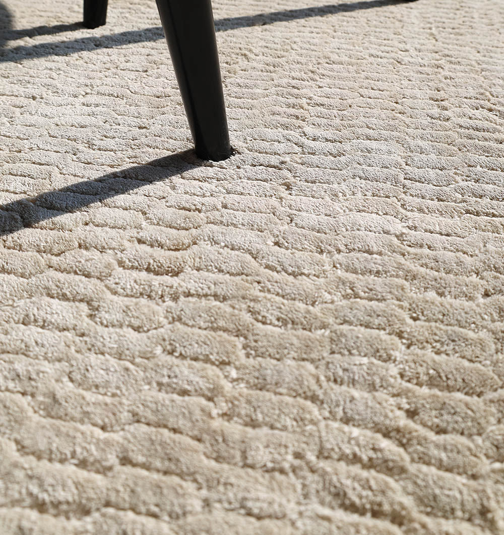 Close-up of the RAMSHA HOME Beige Color Handmade Area Rug - HTV 20 (300x400 cm), showing its subtle wavy pattern with shadows and a black chair leg visible in the upper part of the image.