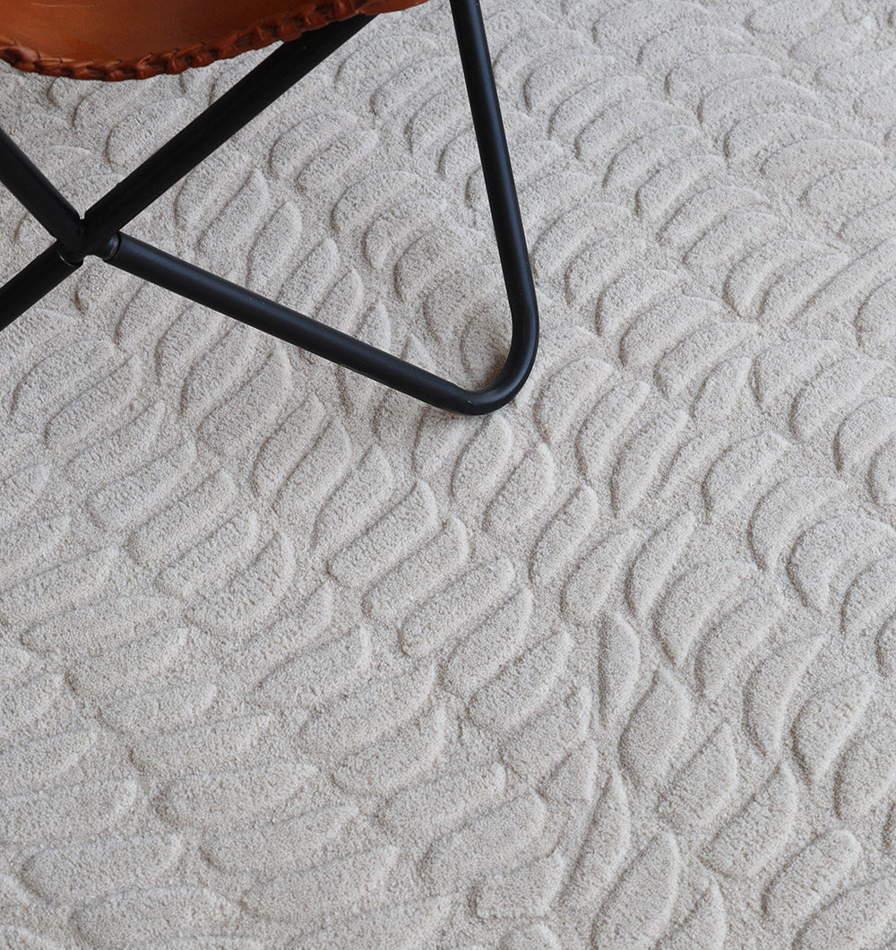 A close-up of the Beige Color Shape Handmade Rug HTW 104 by RAMSHA HOME shows its wavy, embossed texture in light beige. A black metal-framed chair with a brown leather seat appears in the upper left corner.