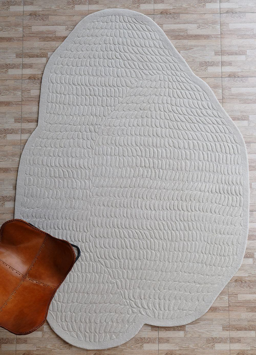 The RAMSHA HOME Beige Color Shape Handmade Rug HTW 104, with its irregular wool design and textured pattern, rests on a light wood floor. A brown leather chair's corner adds to the modern appeal of this hand tufted piece.