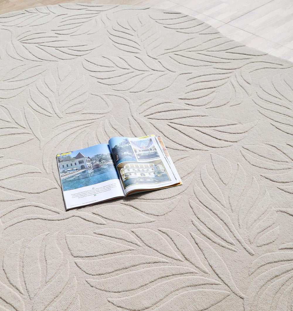An open magazine rests on the Premium Off-White Area Carpet - HTW 115 (200x300 cm) by RAMSHA HOME, its raised leaf pattern adding texture to the wooden floor in a bright, inviting room.