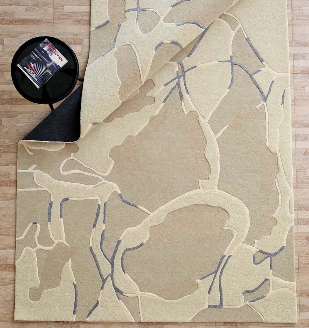 The RAMSHA HOME Abstract Multi-Tone Handmade Carpet – HTWV-175 (200x300 cm) features a modern design in beige and light brown with subtle patterns and a black non-slip backing, shown with a round black table holding a magazine.