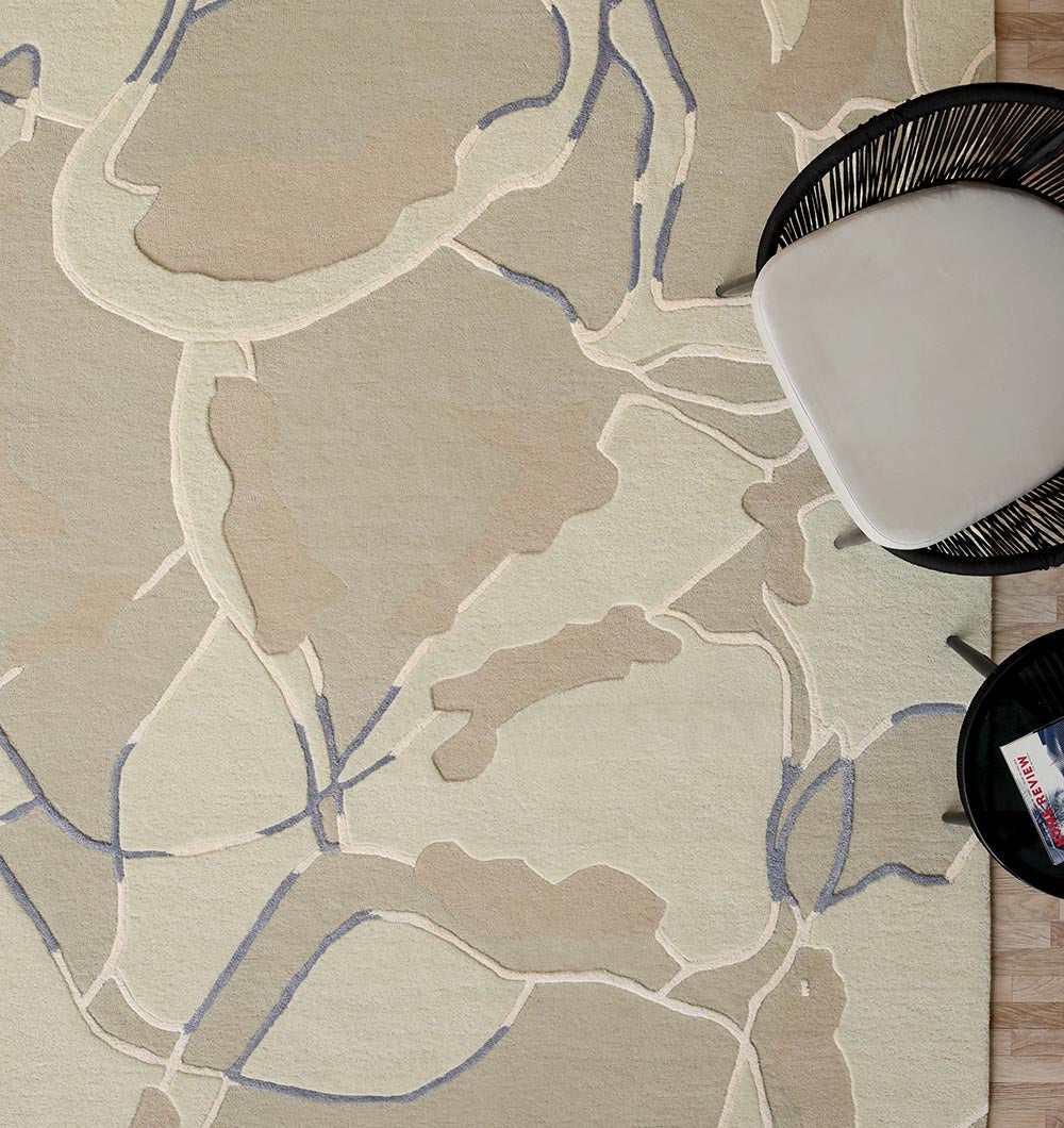 The RAMSHA HOME Abstract Multi-Tone Handmade Carpet – HTWV-175 (200x300 cm) features modern beige, tan, and grey-blue tones, enhancing sophistication beside a black chair and table on the wooden floor.