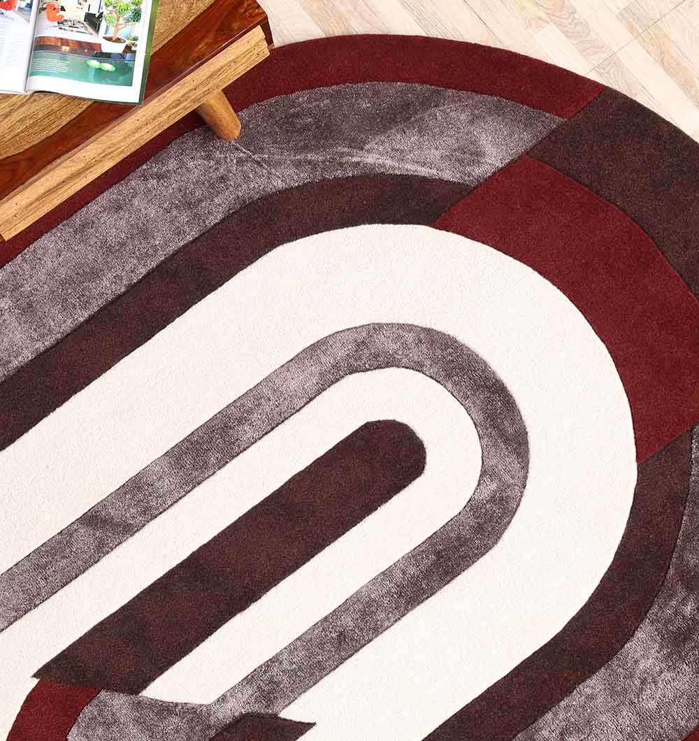 A close-up of the RAMSHA HOME Modern Geometric Shaped Rug HTWV 184 (200 x 226 cm) shows its curved white, brown, and dark red patterns on a light wood floor, with part of a wooden table and magazine in the top left corner.