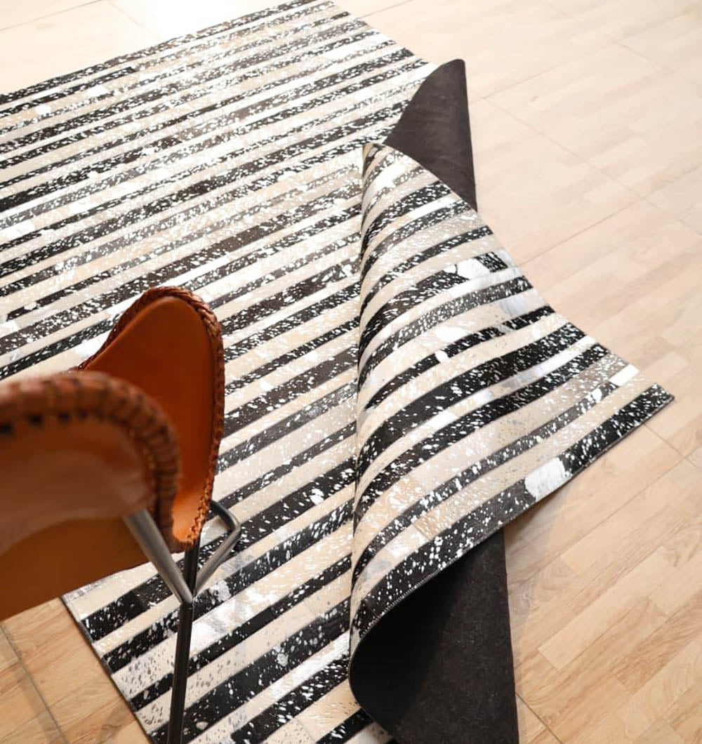 A close-up of the RAMSHA HOME Striped Abstract Black & Ivory Leather Area Rug LR-009, featuring irregular patterns and partially folded, with a tan stitched leather chair on a light wooden floor nearby.