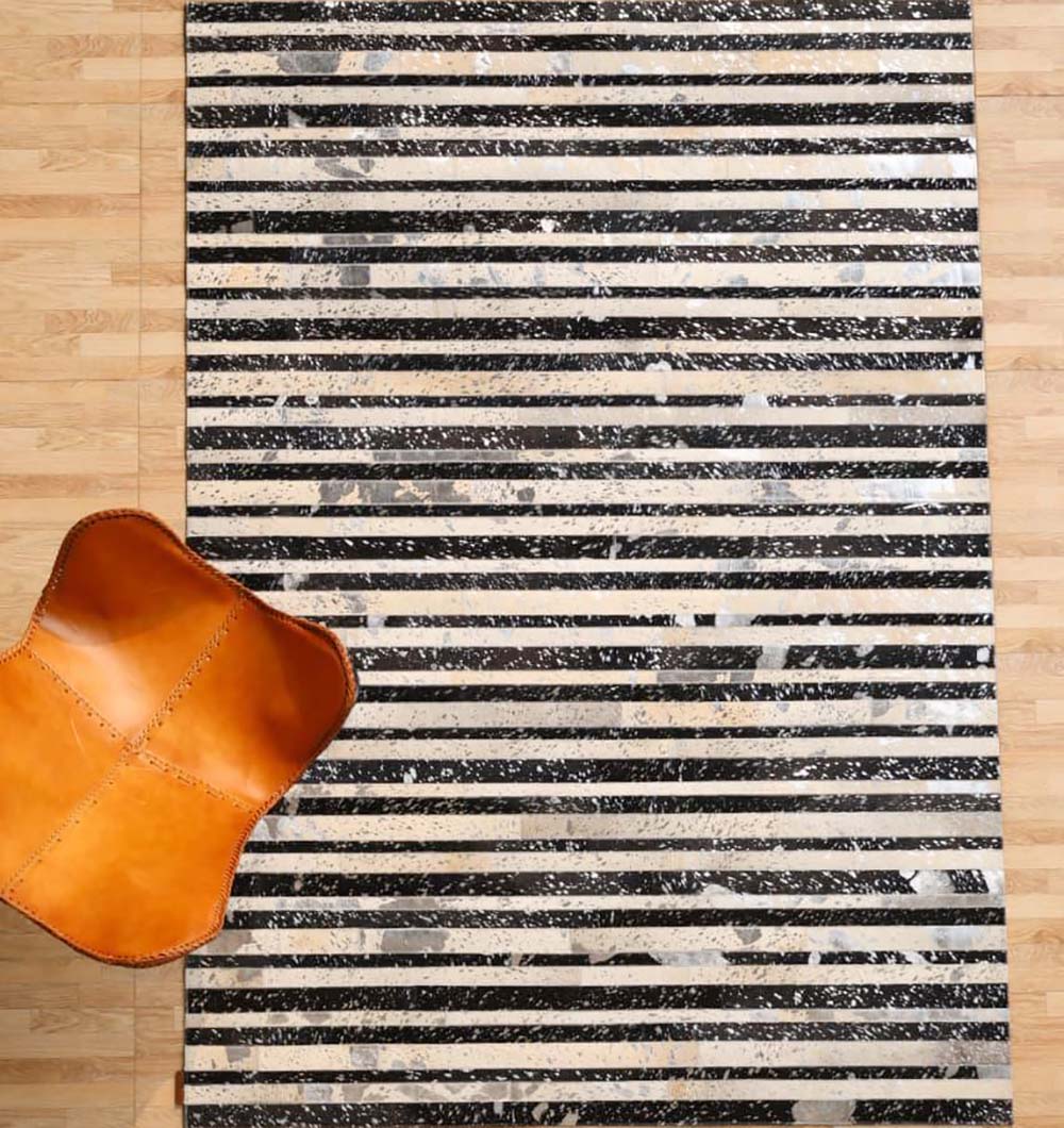 The Striped Abstract Black & Ivory Leather Area Rug LR-009 by RAMSHA HOME is displayed on a light wood floor, with an orange-brown leather chair in the bottom left corner.