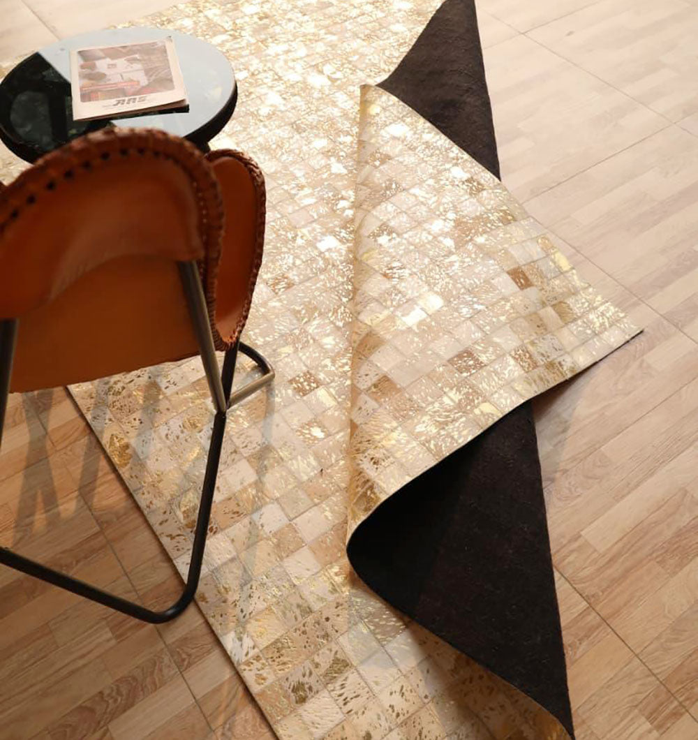 The RAMSHA HOME Handmade Mosaic Patchwork Beige & Tan Leather Rug LR-015 is partially flipped over on a wooden floor next to a brown leather chair and a small black round table with a magazine, sunlight streaming in from the right.