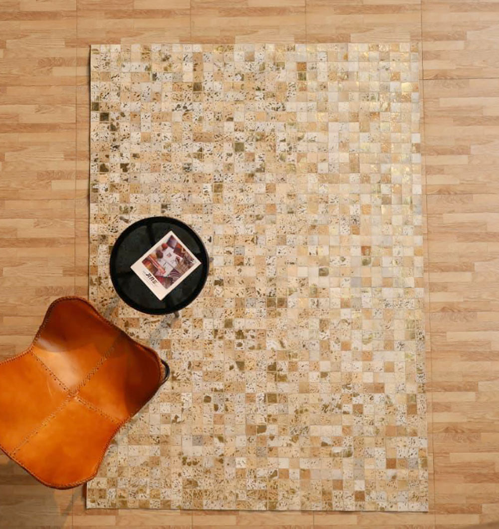 A top view of a tan leather chair, a round black side table with a magazine, and the RAMSHA HOME Handmade Mosaic Patchwork Beige & Tan Leather Rug LR-015 on a light wood floor.