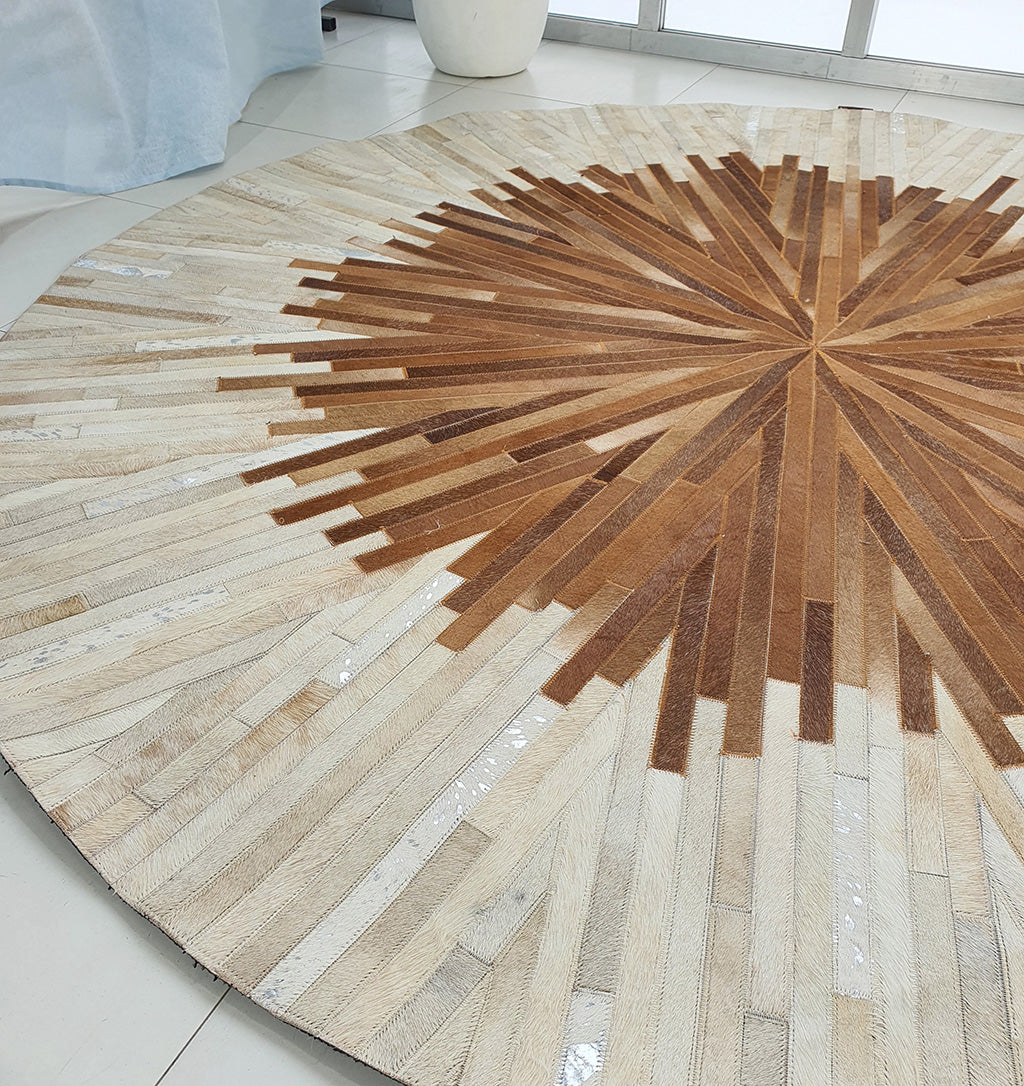 The Multi Color Round Leather Rug - LR 016 features a geometric sunburst pattern in brown and beige, displayed on a light tile floor beside large windows and a white planter.