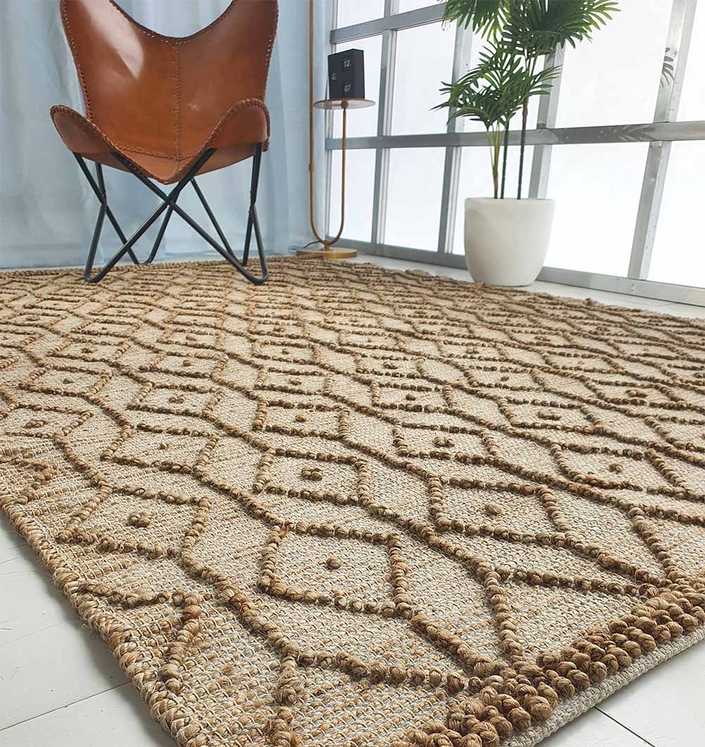 A close-up of the RAMSHA HOME Brown Rectangle Area Rug and Carpet for Stylish Living Room SR-004, featuring a hand-knotted diamond pattern, placed on a light floor beside a brown leather chair, potted plant, and sunlit windows.
