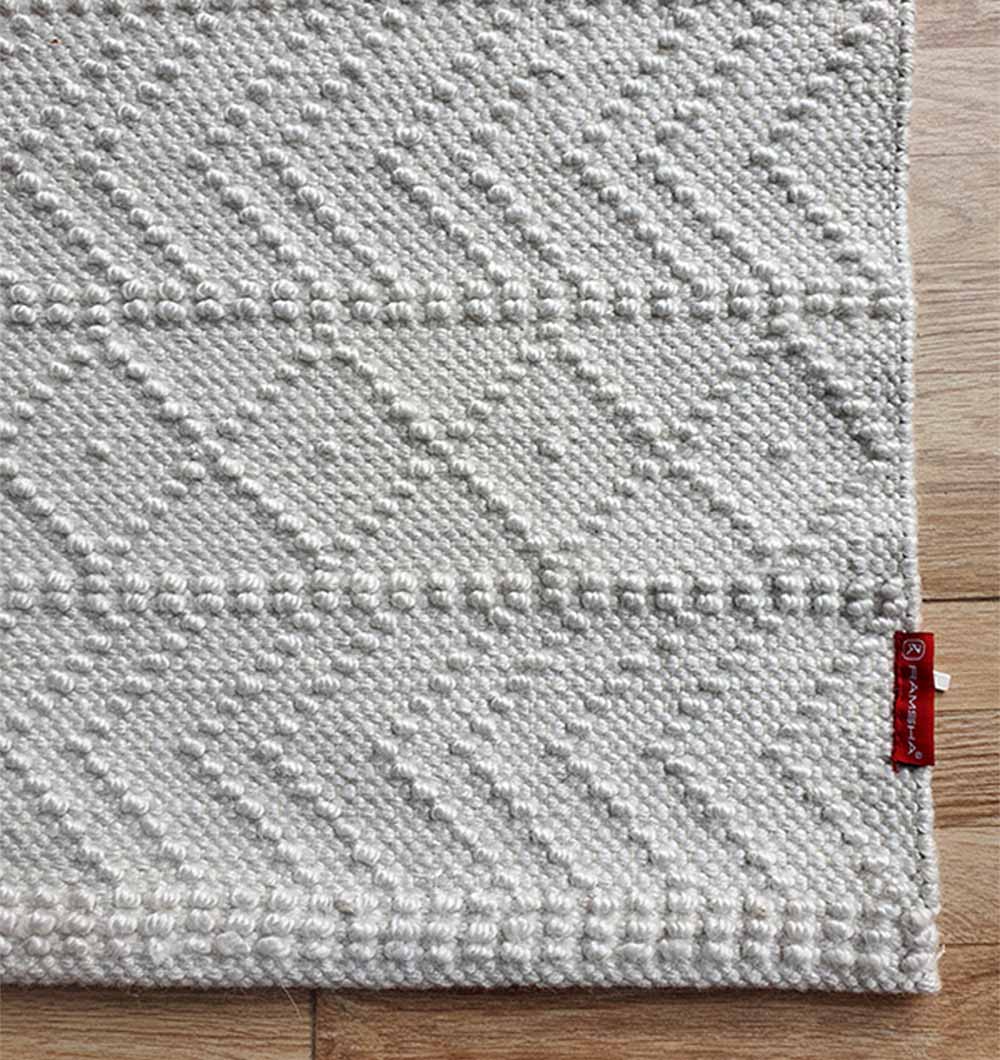 A close-up of the RAMSHA HOME White Rectangle Woven Area Rug SR-022, featuring geometric patterns on a durable polyester surface with a red fabric tag, displayed on a wooden floor in a living room or bedroom.