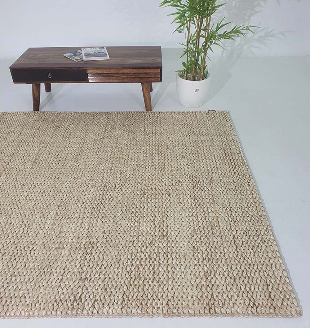 The RAMSHA HOME Bleach Color Woven Floor Rug For Home Decor SR-037 in textured beige lies on a white floor, paired with a dark wooden coffee table and a potted green plant in the background.