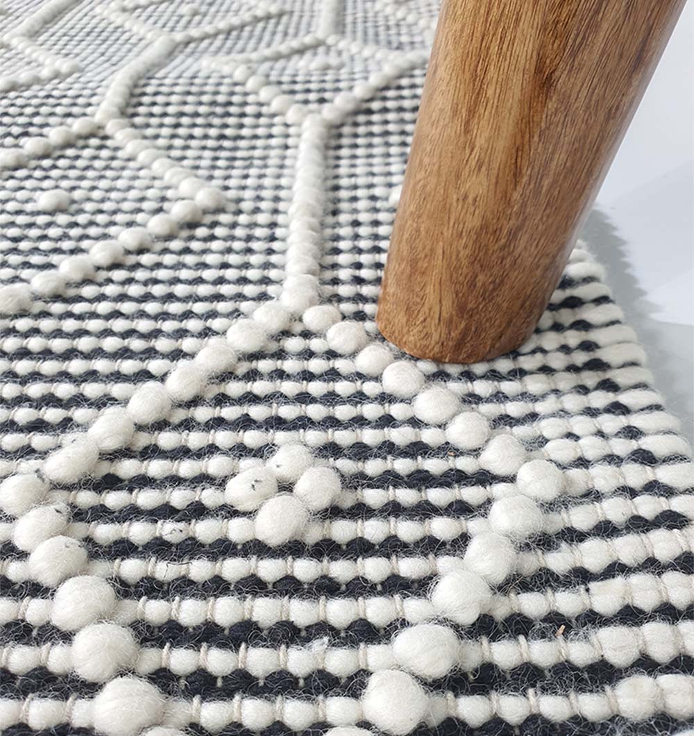 A close-up of a wooden furniture leg on the RAMSHA HOME Hand Woven Fringes Rug SRN-018 in natural and black, featuring a geometric woven design with raised, tufted dots.
