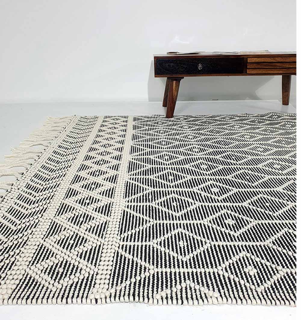 The RAMSHA HOME Hand Woven Fringes Rug Natural & Black Color SRN-018 with bold geometric patterns lies on a light floor. A wooden table with angled legs and drawers appears in the background against a white wall.