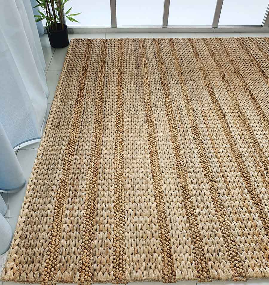 The RAMSHA HOME Natural Color Hand Woven Jute Rug SR-043, featuring beige and brown stripes, lies on a light floor by a window with sheer curtains and a potted plant in the corner.