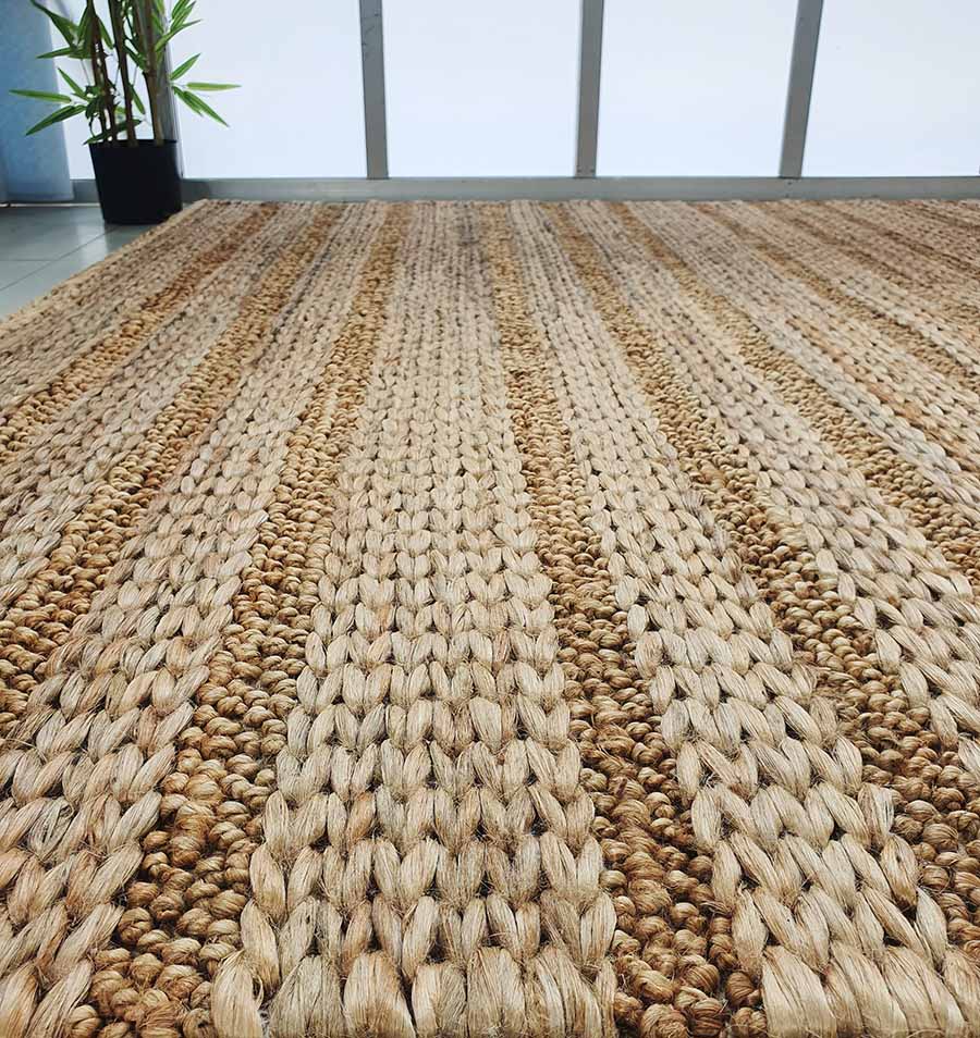 Close-up of the RAMSHA HOME Natural Color Hand Woven Jute Rug SR-043, featuring thick braided patterns in natural tones. Placed near large windows with a potted plant, it's an ideal Ramadan gift made from natural fibers.