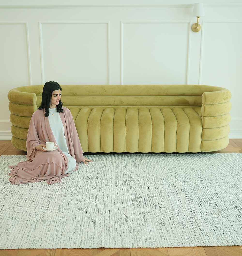 A woman in a pink robe sits on the Durable Handmade Natural White Woven Area Rug SR-058 by RAMSHA HOME, holding a cup and saucer in front of a modern mustard-yellow sofa against a white paneled wall with a gold sconce.